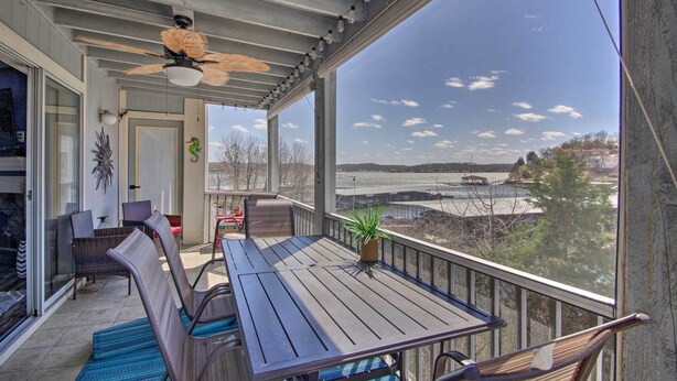 A covered outdoor area with a table and chairs, a ceiling fan, and a view of a body of water and distant landscape. - Lake-view Patio, Pool Access! Regatta Bay Condo (Lake Ozark)