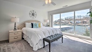 A bedroom with a large window, a ceiling fan, a bed with white bedding, a bedside table with lamps, and a view of a residential area. - Lake-view Patio, Pool Access! Regatta Bay Condo (Lake Ozark)