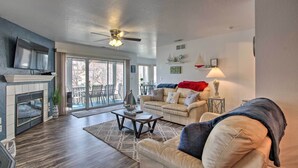 A spacious living room with a fireplace, a ceiling fan, a television, and a large window overlooking a deck. - Lake-view Patio, Pool Access! Regatta Bay Condo (Lake Ozark)