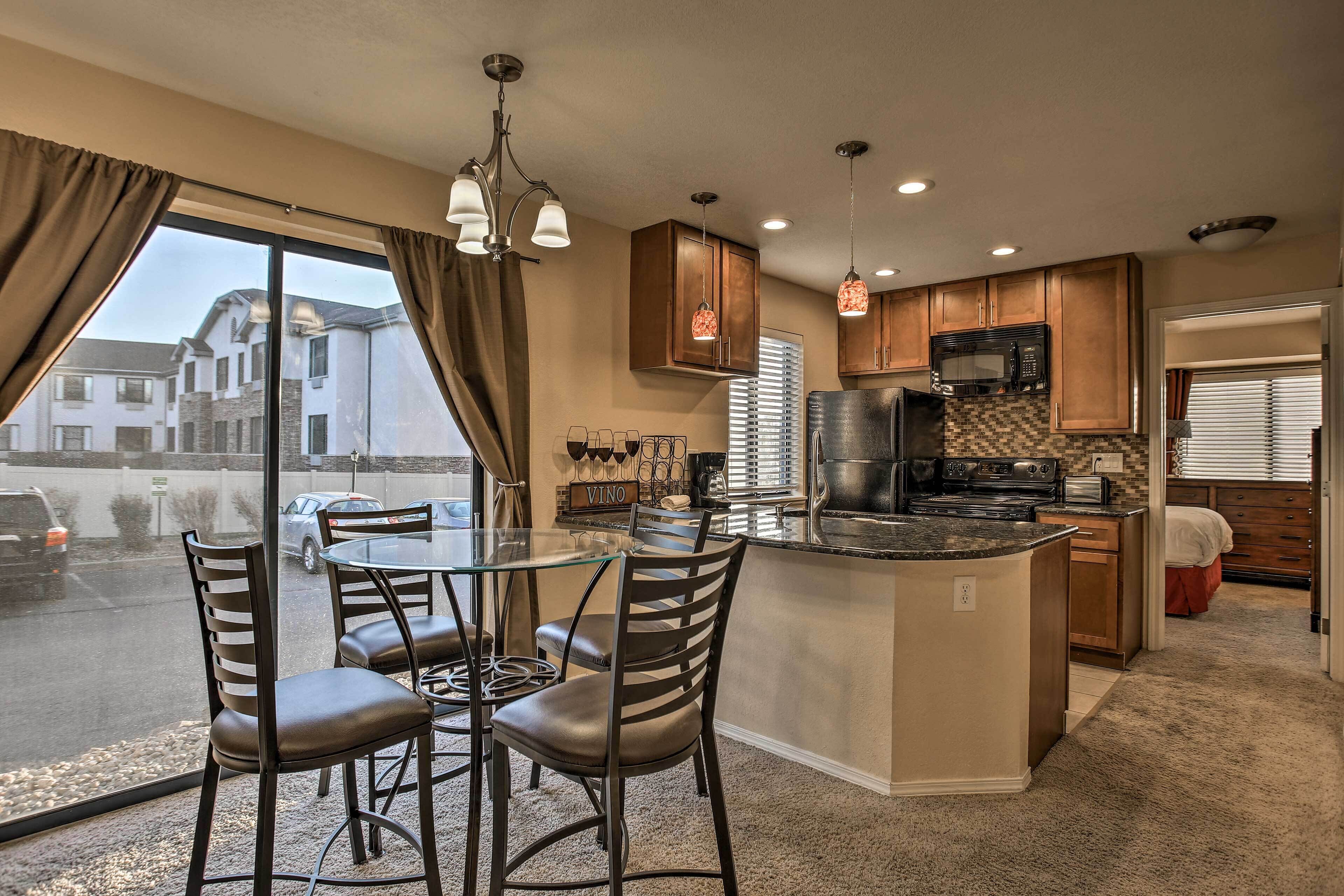 A modern kitchen with a central island, dining area with chairs, and large windows offering a view of the street.