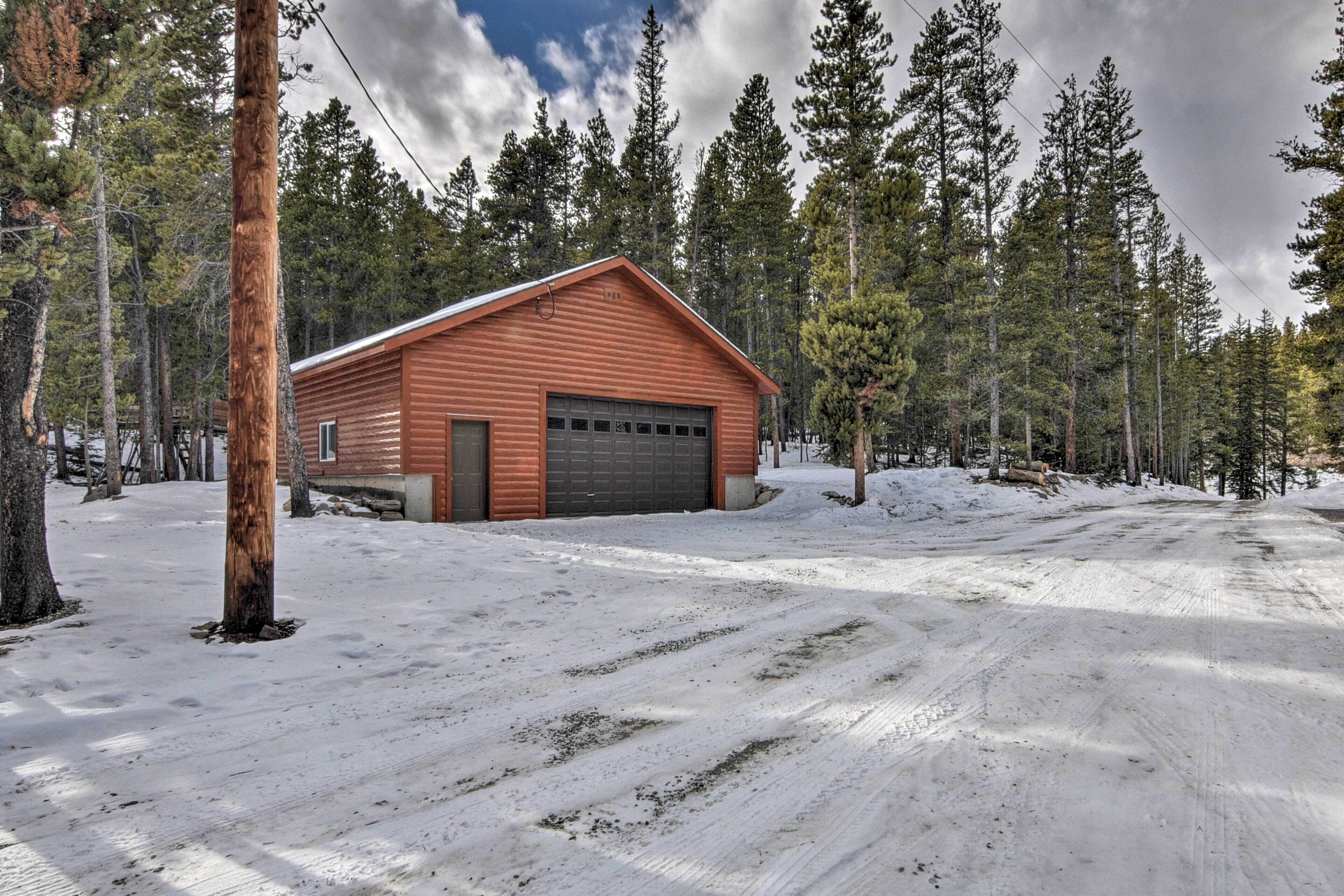 A wooden cabin with a garage, surrounded by tall pine trees and snow-covered ground.