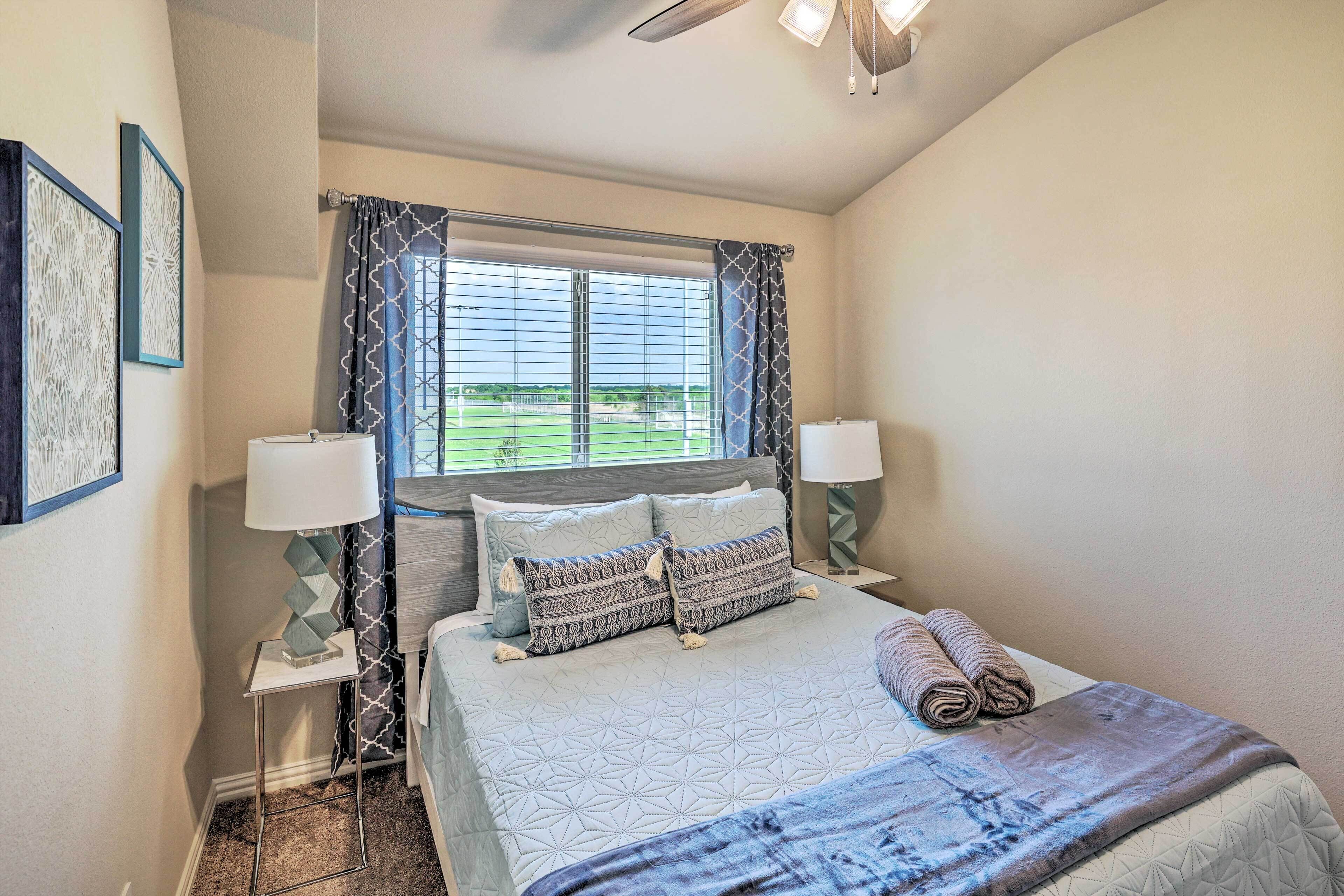 A bedroom with a bed, two bedside lamps, a window with curtains, and a view of a green landscape.