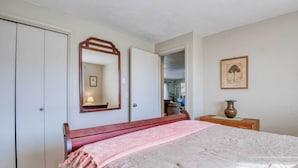 Un dormitorio con una cama, cabecera de madera, mesita de noche con un florero, un espejo y una puerta que da a otra habitación.