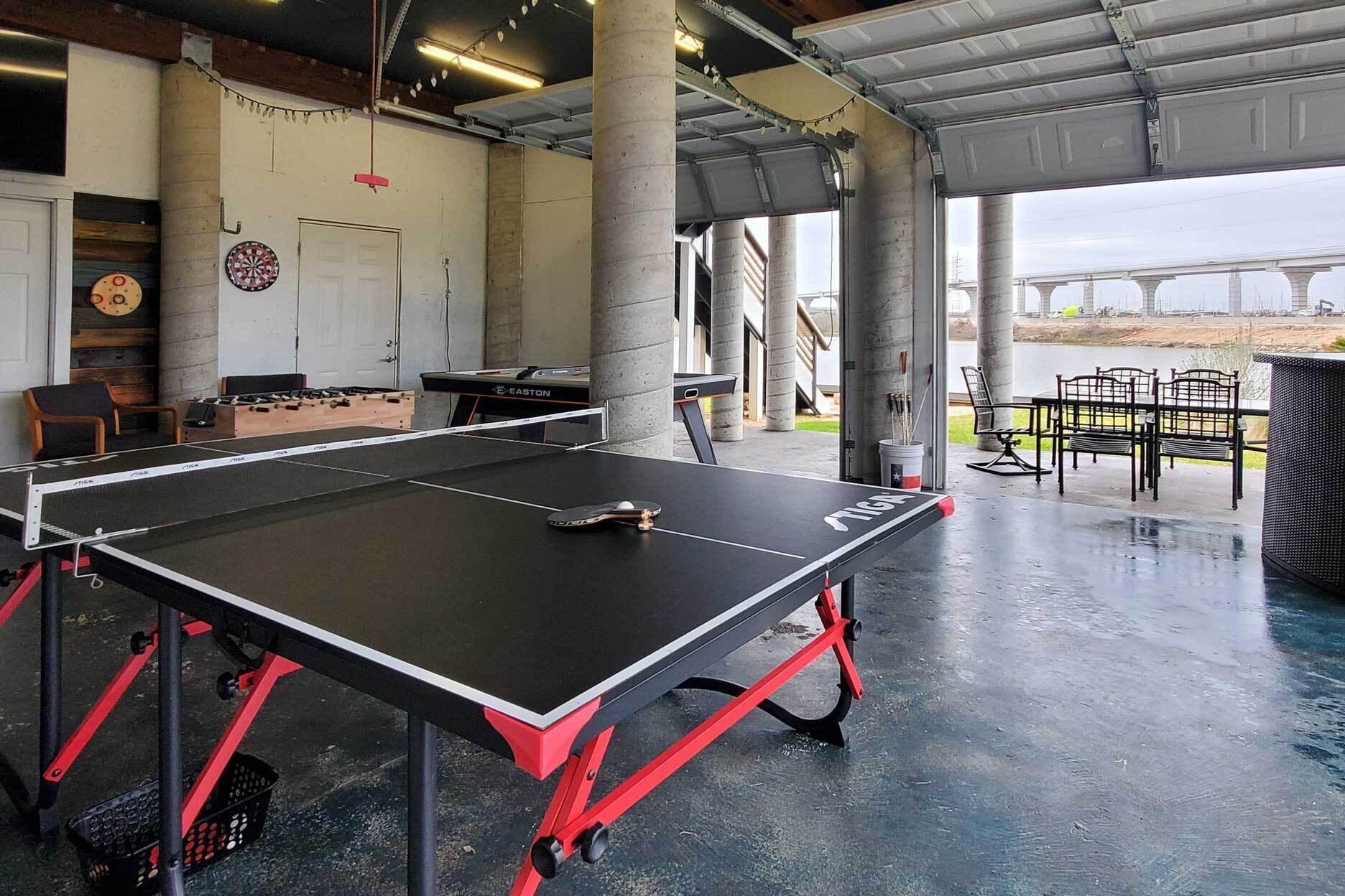 A ping pong table in a covered area with concrete pillars, a dartboard, and a foosball table.