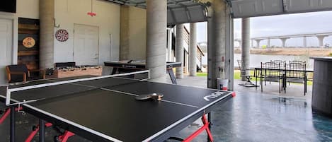 A ping pong table in a covered area with concrete pillars, a dartboard, and a foosball table.