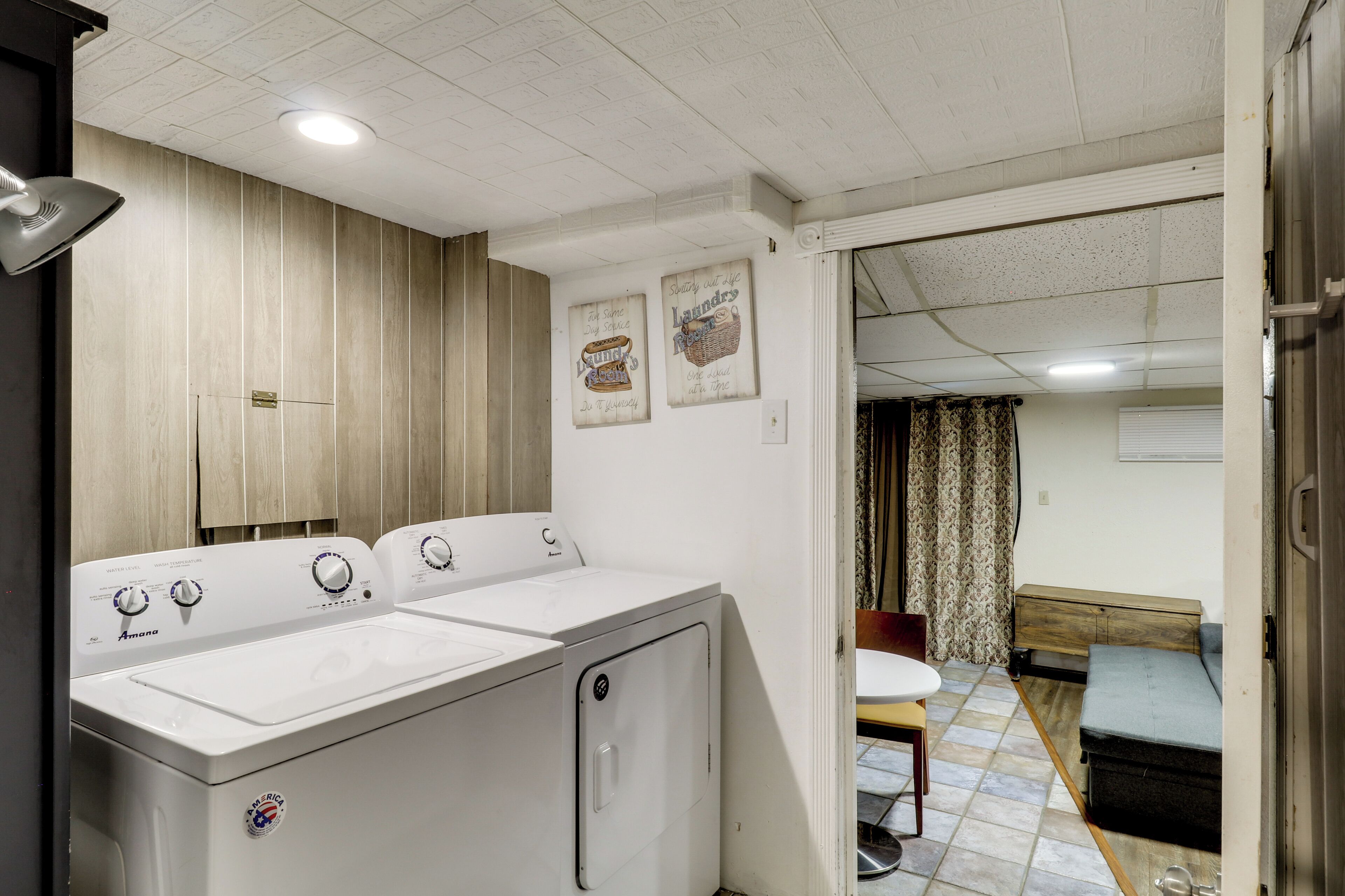 Un cuarto de lavandería con dos lavadoras blancas, paneles de madera en las paredes y un comedor visible a través de una puerta abierta.