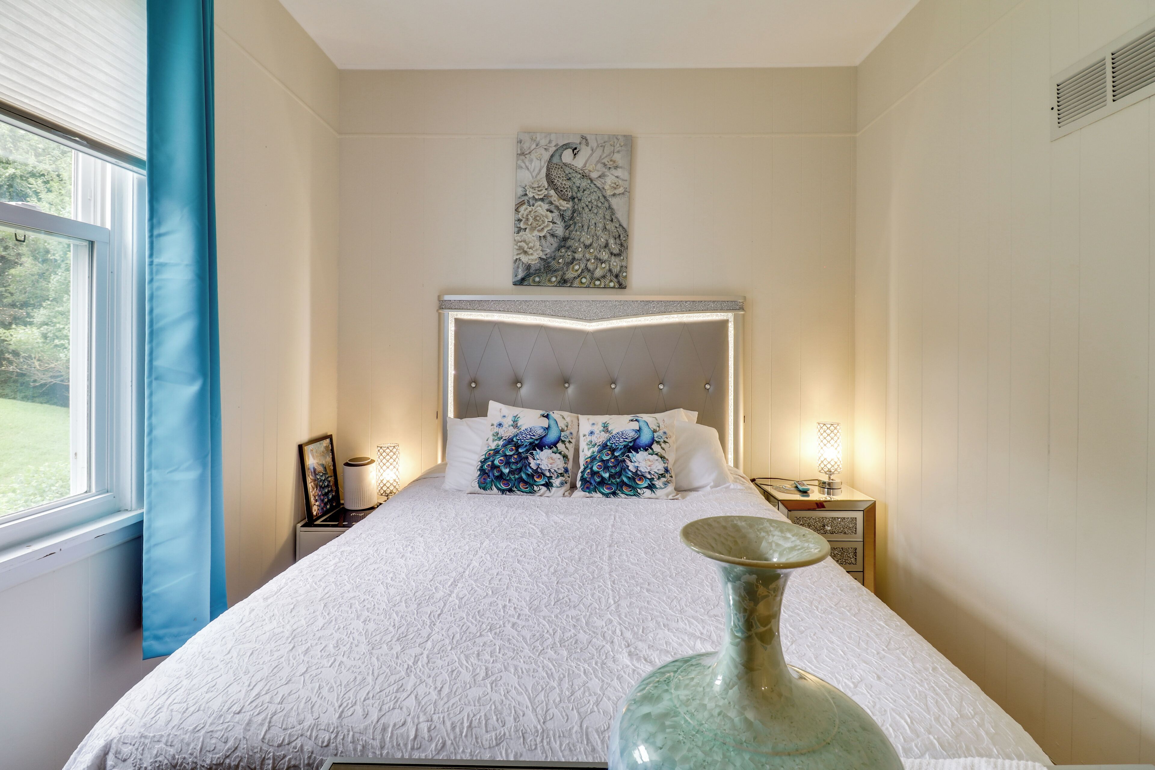 Un dormitorio con una cama grande, un jarrón decorativo, un cuadro sobre la cama y una ventana con cortinas.