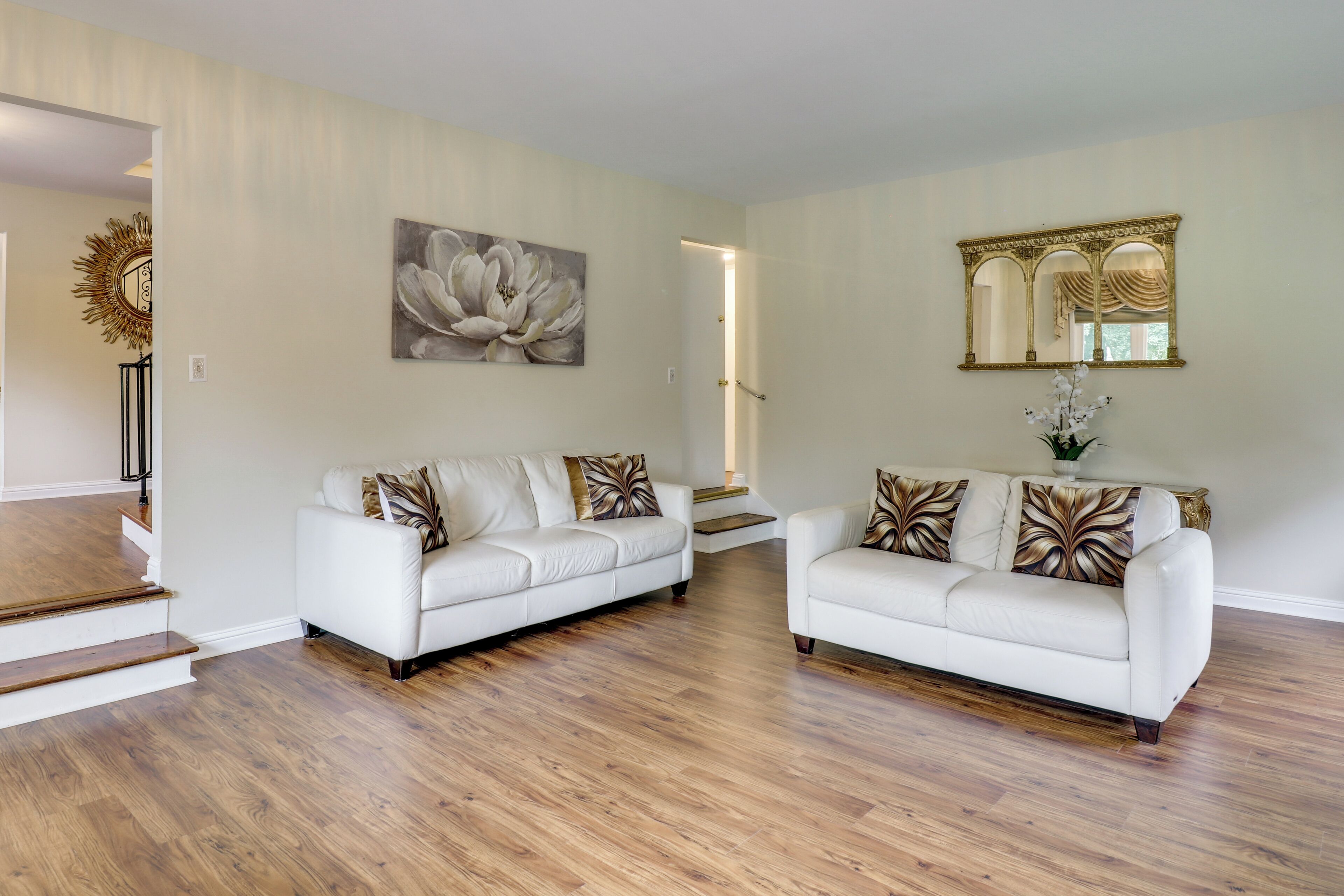 A spacious living area with two white sofas, a large floral painting, and a mirror with an ornate frame.