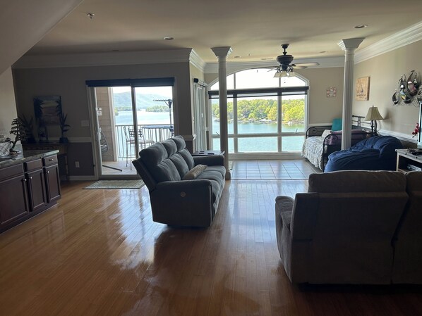 A spacious living room with a large window offering a view of a lake and mountains. There is a dining area with a table and chairs, and a kitchen with dark wooden cabinets.