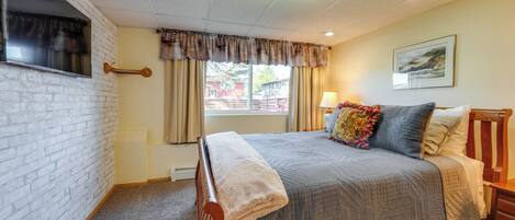 A bedroom with a bed, a nightstand, a window with curtains, a wall-mounted TV, and a picture on the wall.