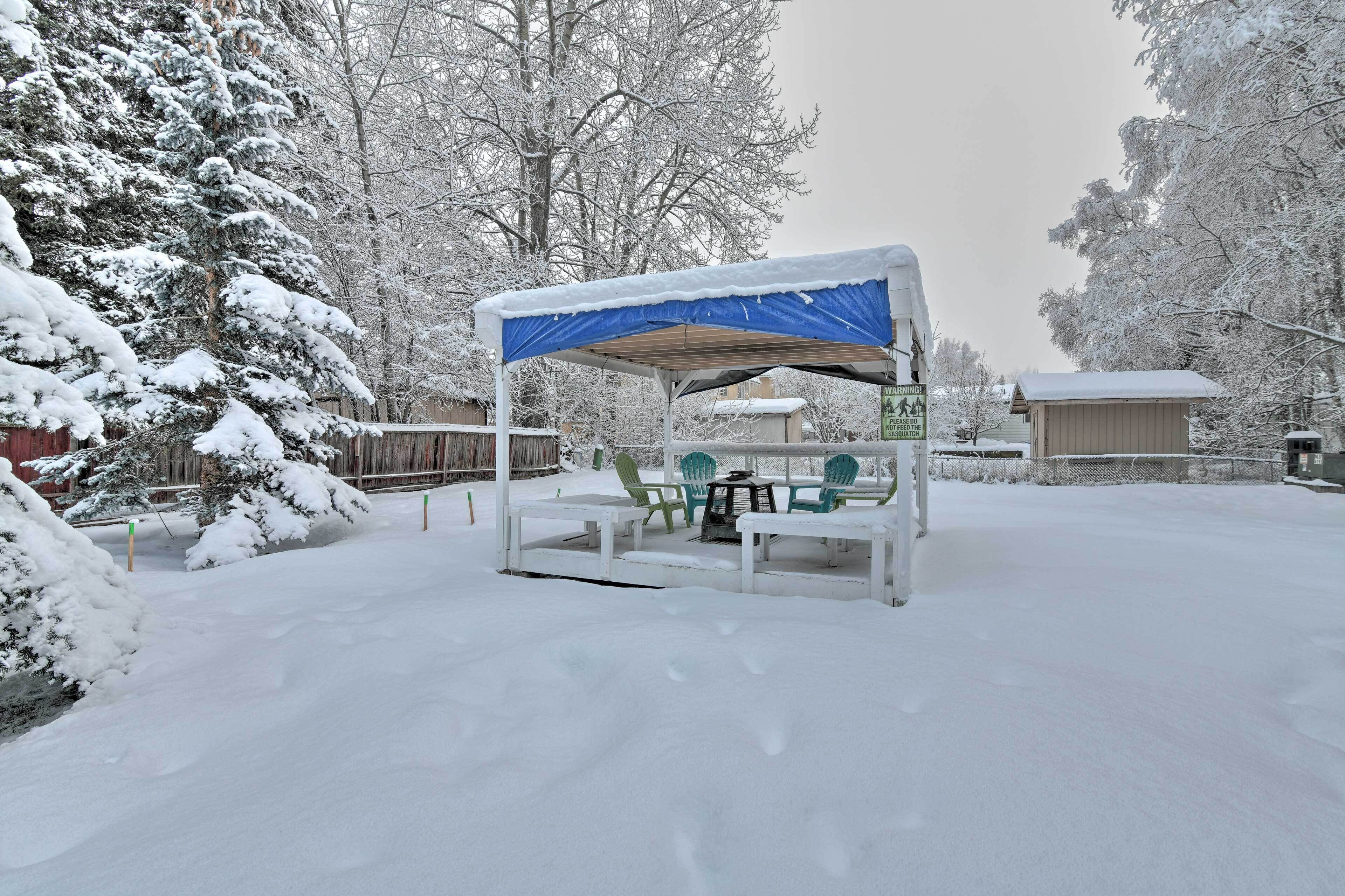 Een overdekt terras met een tafel en stoelen, omringd door met sneeuw bedekte bomen en een houten hek.