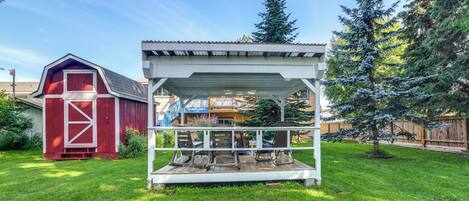 A covered outdoor seating area with a white roof and wooden deck, surrounded by a well-maintained lawn and a red barn in the background.