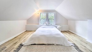 Een slaapkamer met een schuine kap, een raam met uitzicht op groen, een bed met een witte sprei en een nachtkastje.