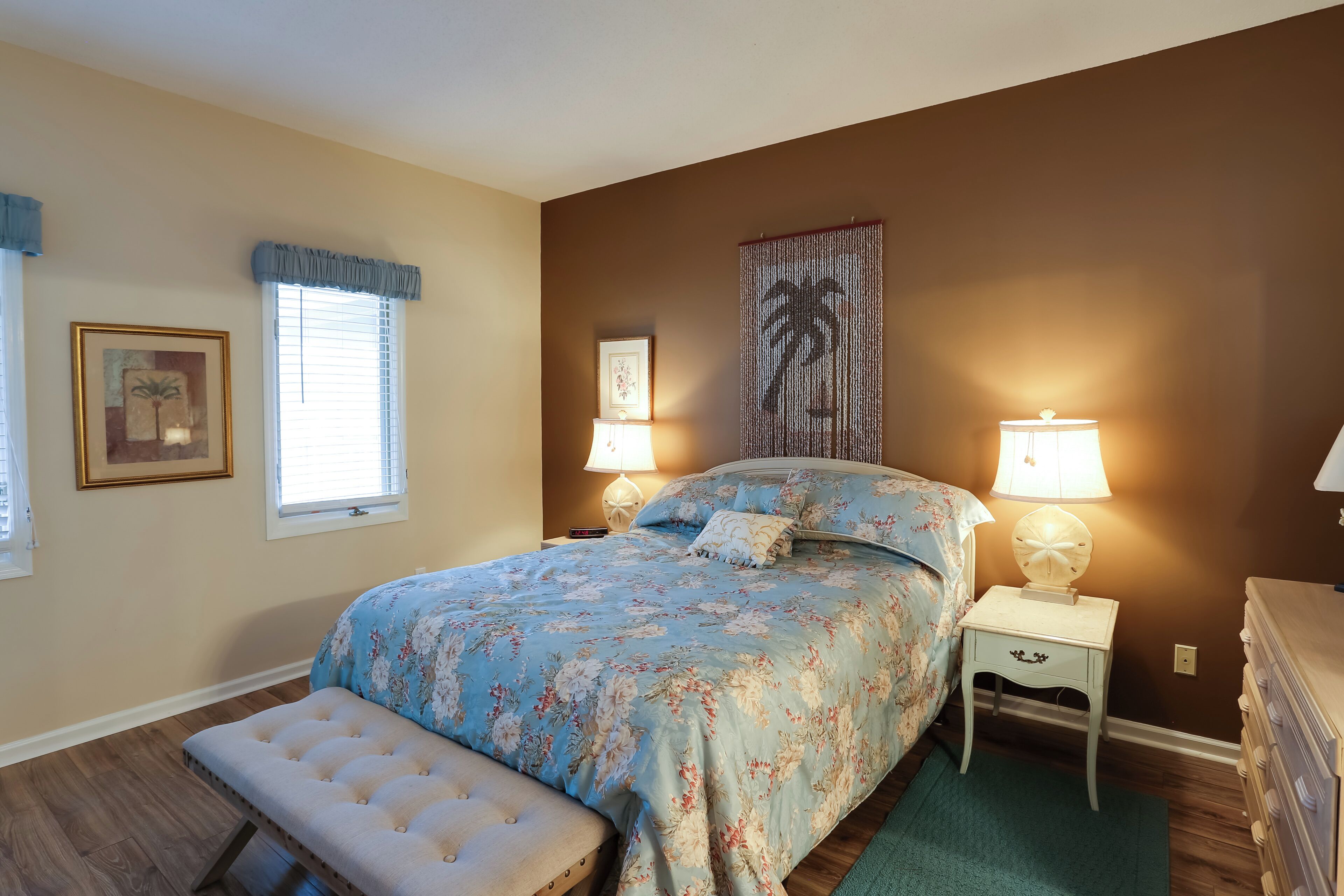 A bedroom with a floral bedspread, a bench, two bedside lamps, a wall art piece, and a dresser.