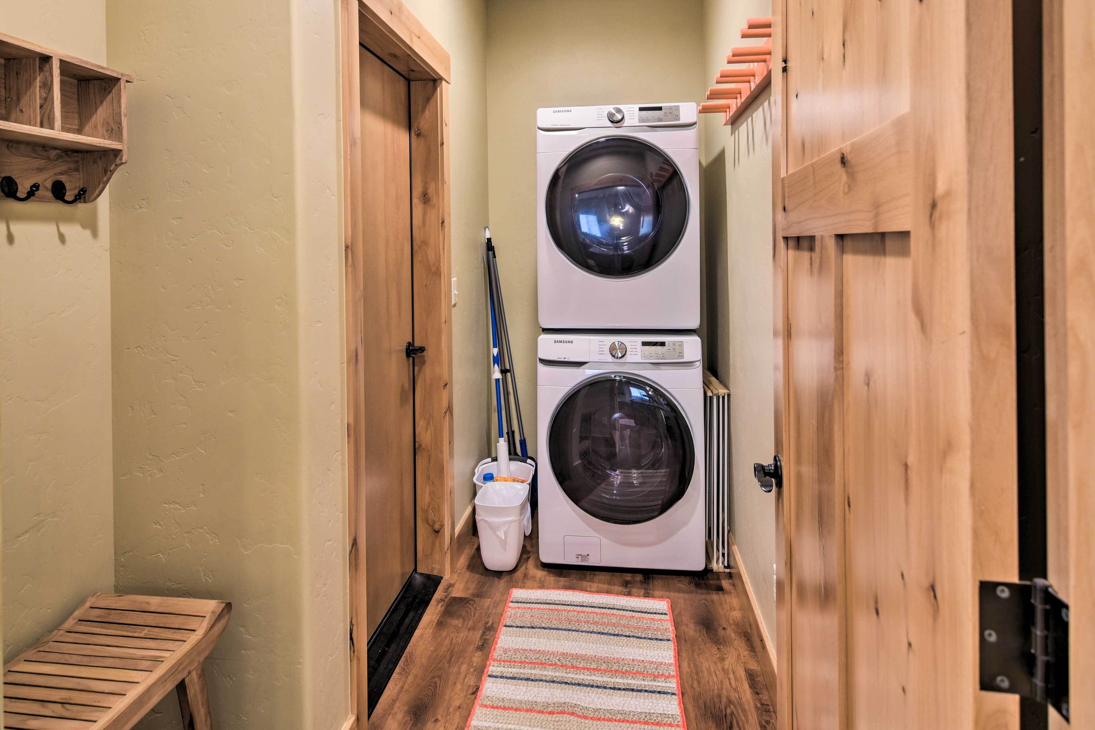 A laundry room with two stacked front-loading washing machines, a wooden bench, a striped rug, and a mop leaning against the wall.
