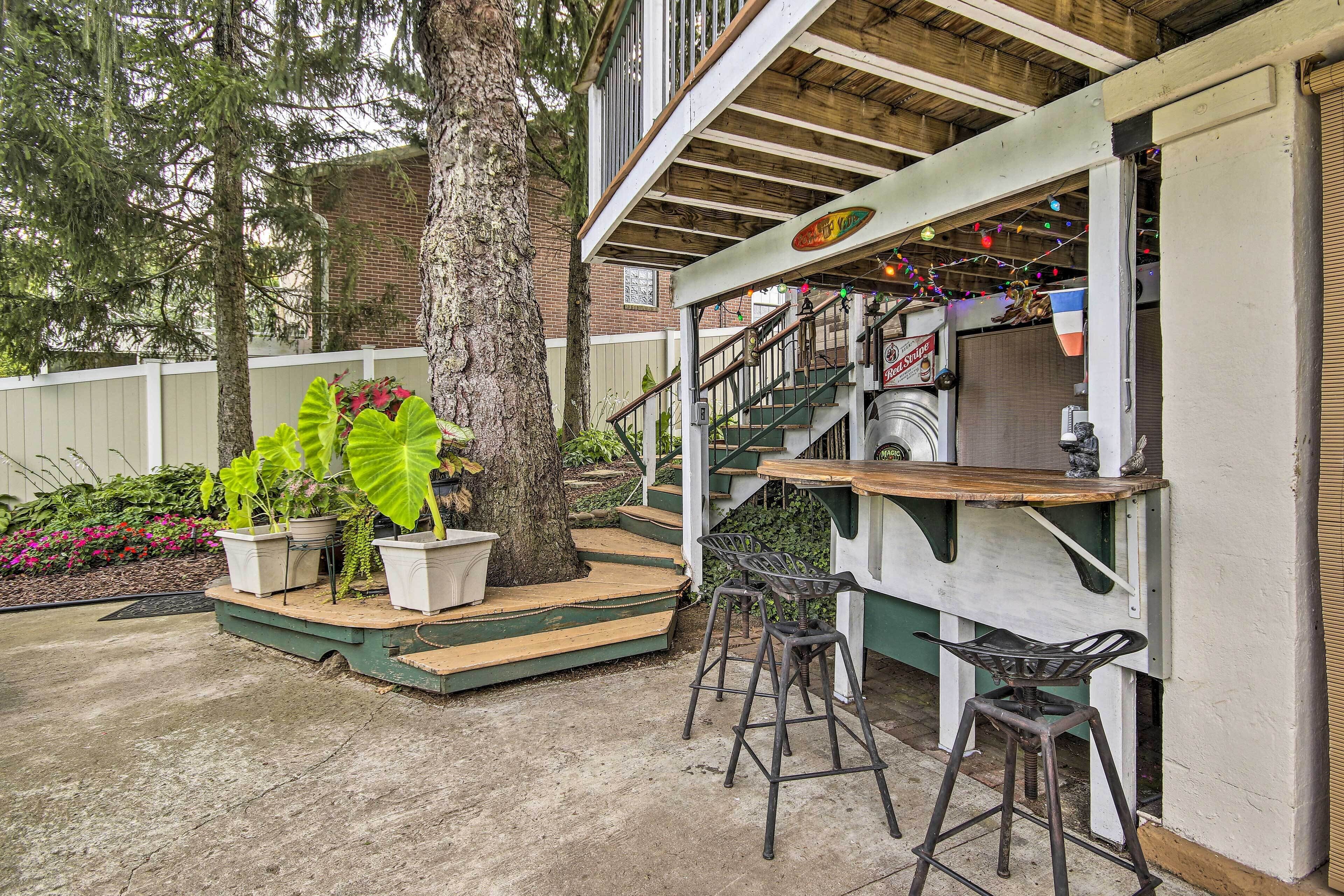 Un patio al aire libre con piso de madera, dos taburetes de metal, una mesita y una escalera que sube a un nivel superior.