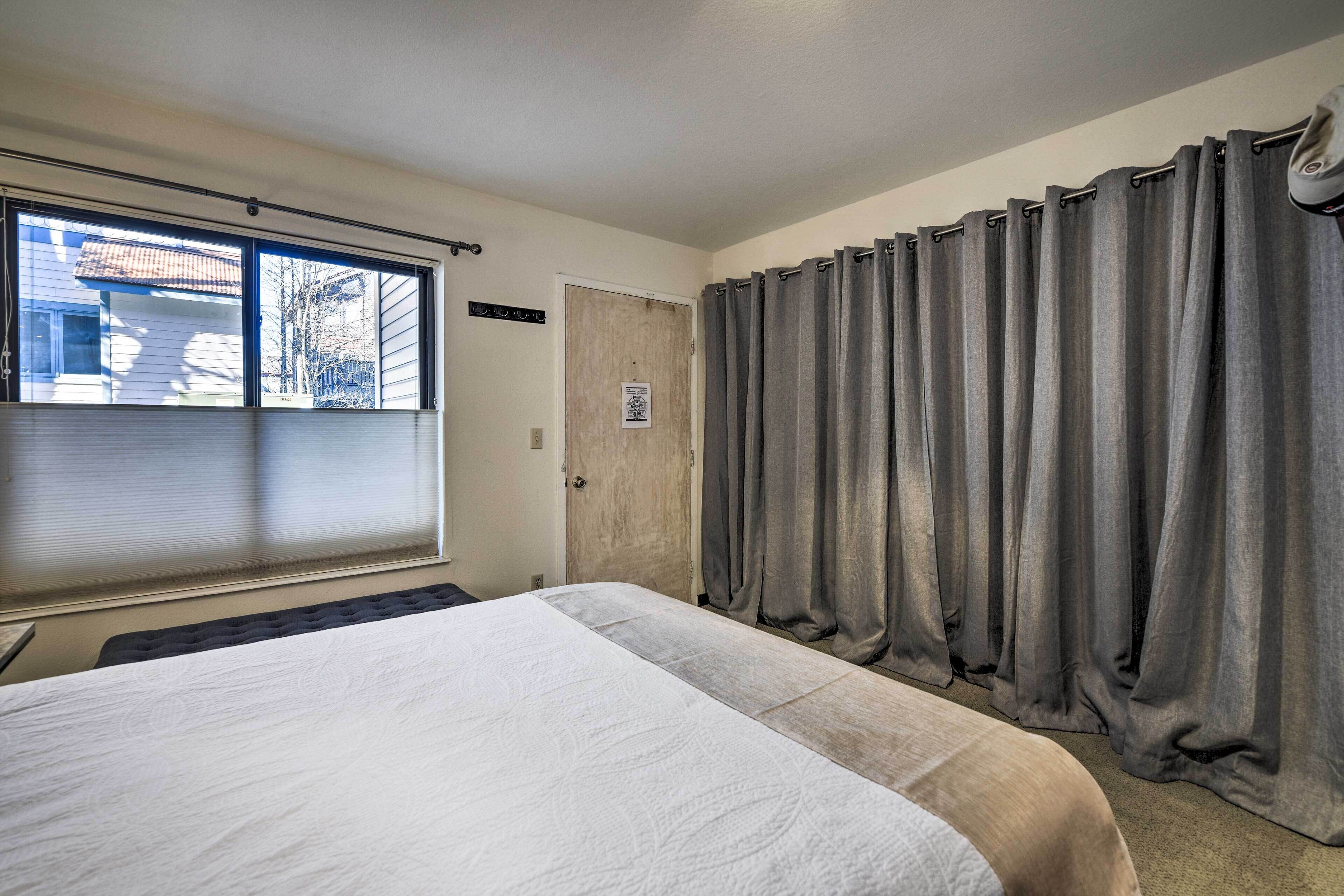 A bedroom with a large bed, a window with blinds, a door, and grey curtains.