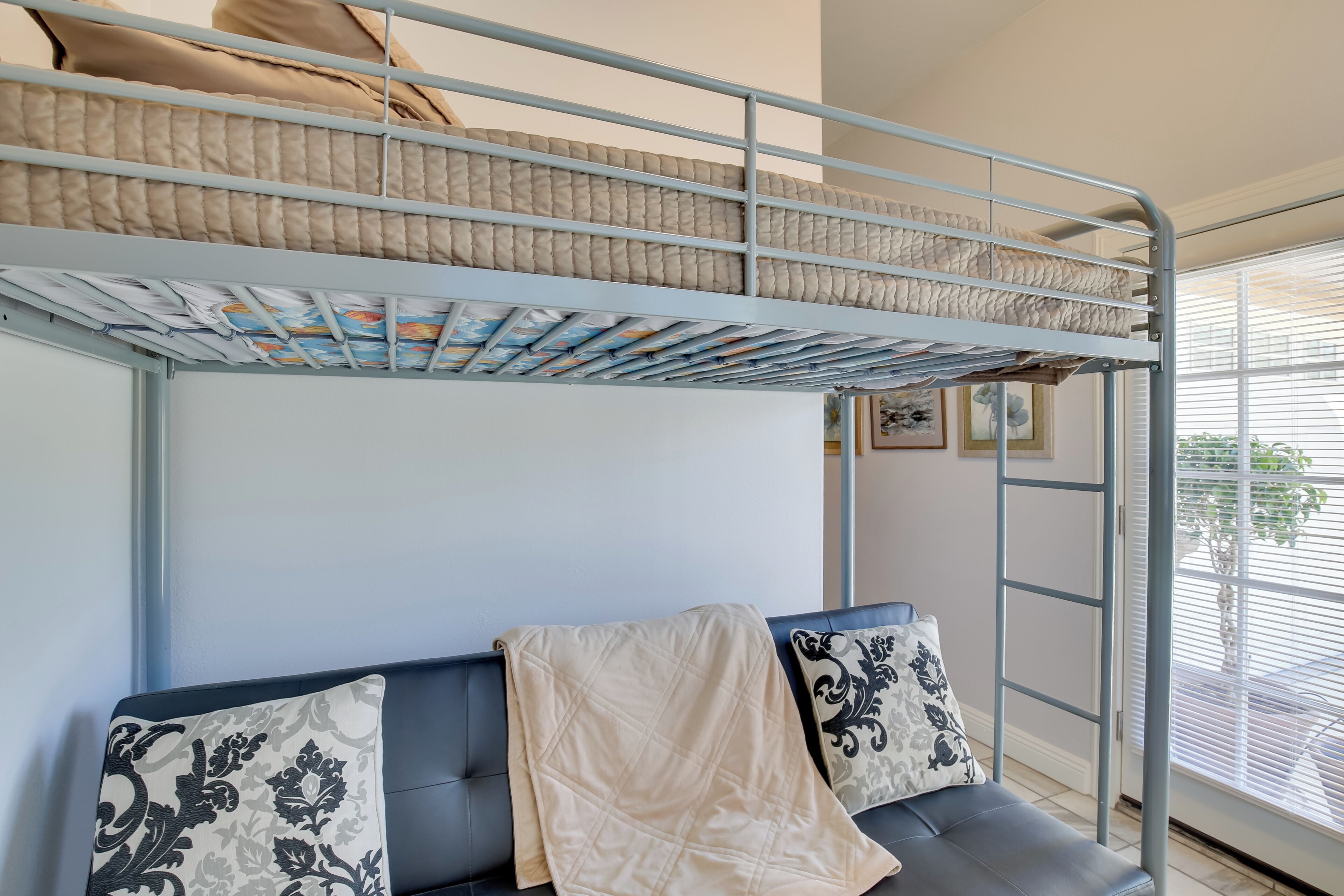 A bunk bed with a quilted mattress, a sofa with patterned cushions, and a window with blinds.