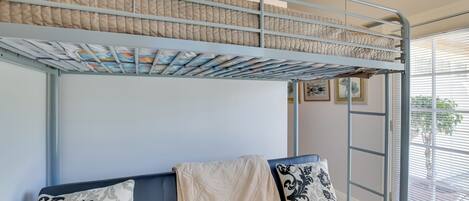 A bunk bed with a quilted mattress, a sofa with patterned cushions, and a window with blinds.