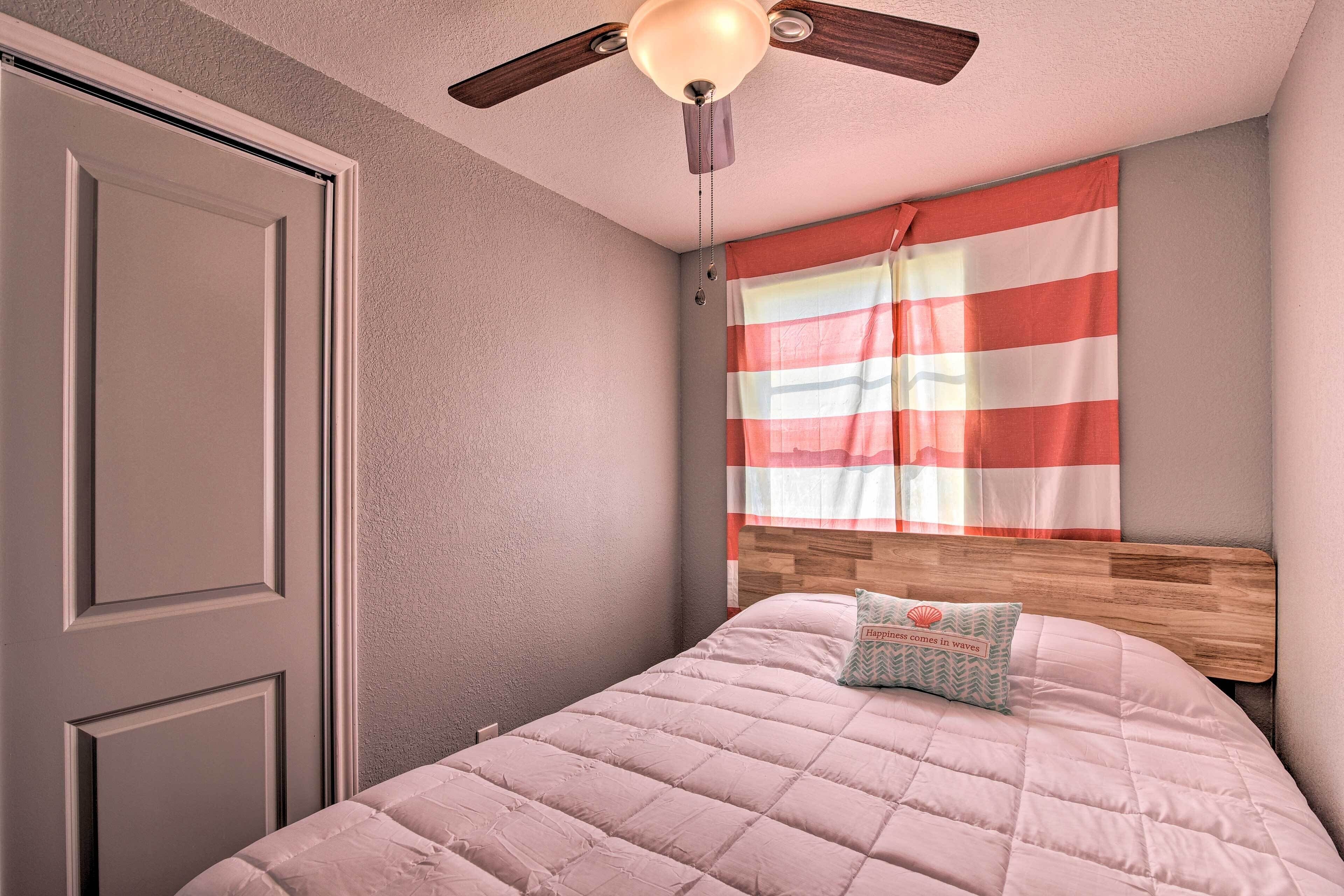 A bedroom with a bed, a ceiling fan, a window with striped curtains, and a wooden headboard.