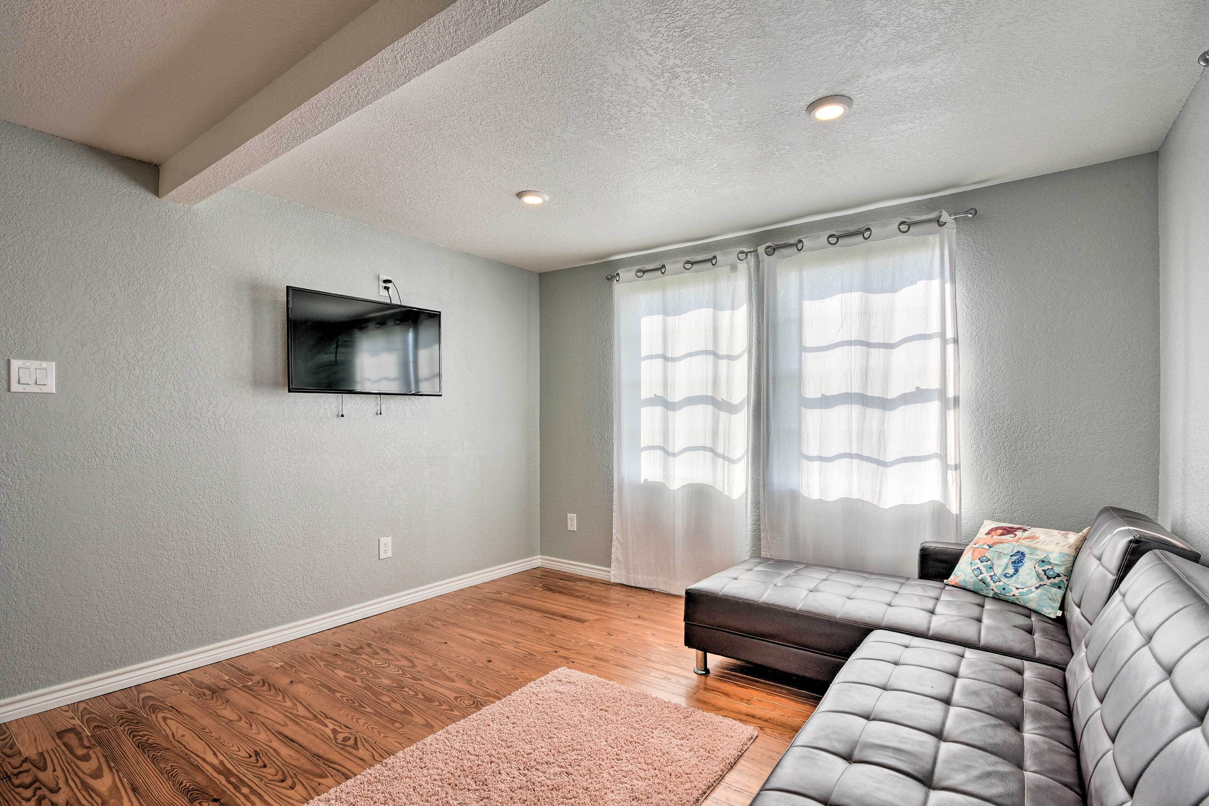 A living room with a sectional sofa, a flat-screen TV mounted on the wall, and a large window with sheer curtains.