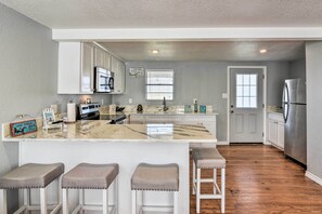 A kitchen with white cabinets, a marble countertop, and a stainless steel refrigerator. There are three stools with cushioned seats and a window with blinds. - 4 Blocks to Ocean: Water-view Surfside Beach Home! (Freeport)