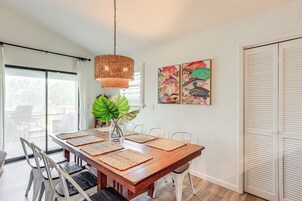 Un comedor con una mesa de madera, sillas blancas, individuales tejidas, una lámpara colgante y una planta verde grande.