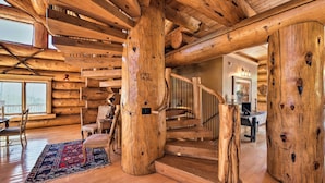 A log cabin interior with a wooden staircase, rustic wooden beams, and a large window.