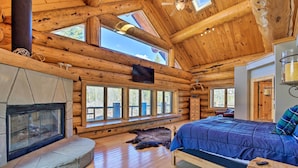 A log cabin interior with a stone fireplace, a large bed, and multiple windows offering views of the outdoors.
