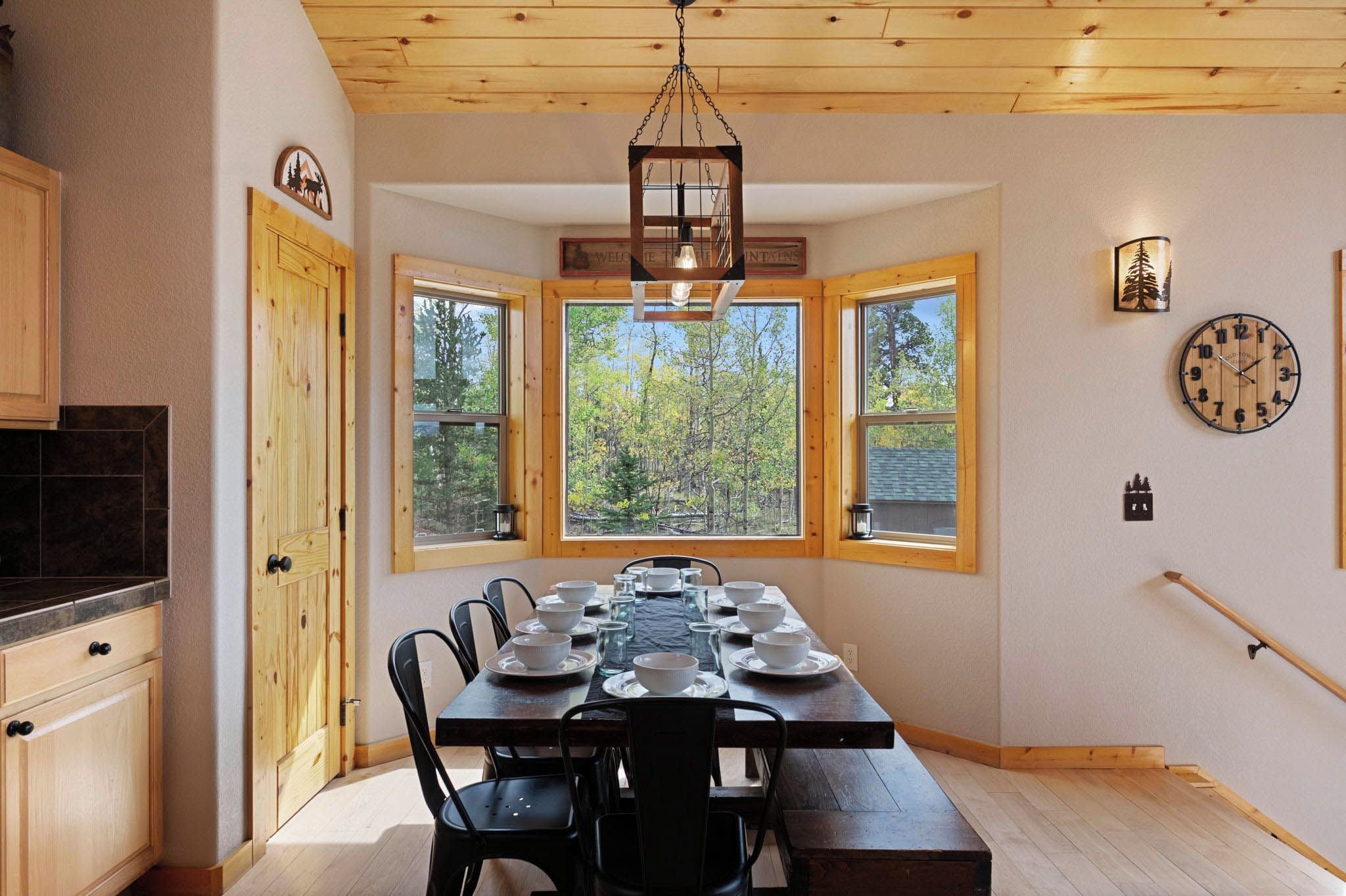 A dining area with a wooden table set for four, surrounded by chairs, with a view of trees through large windows.