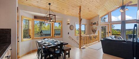 A spacious dining area with a wooden table and chairs, large windows offering views of the outdoors, and a wooden ceiling.