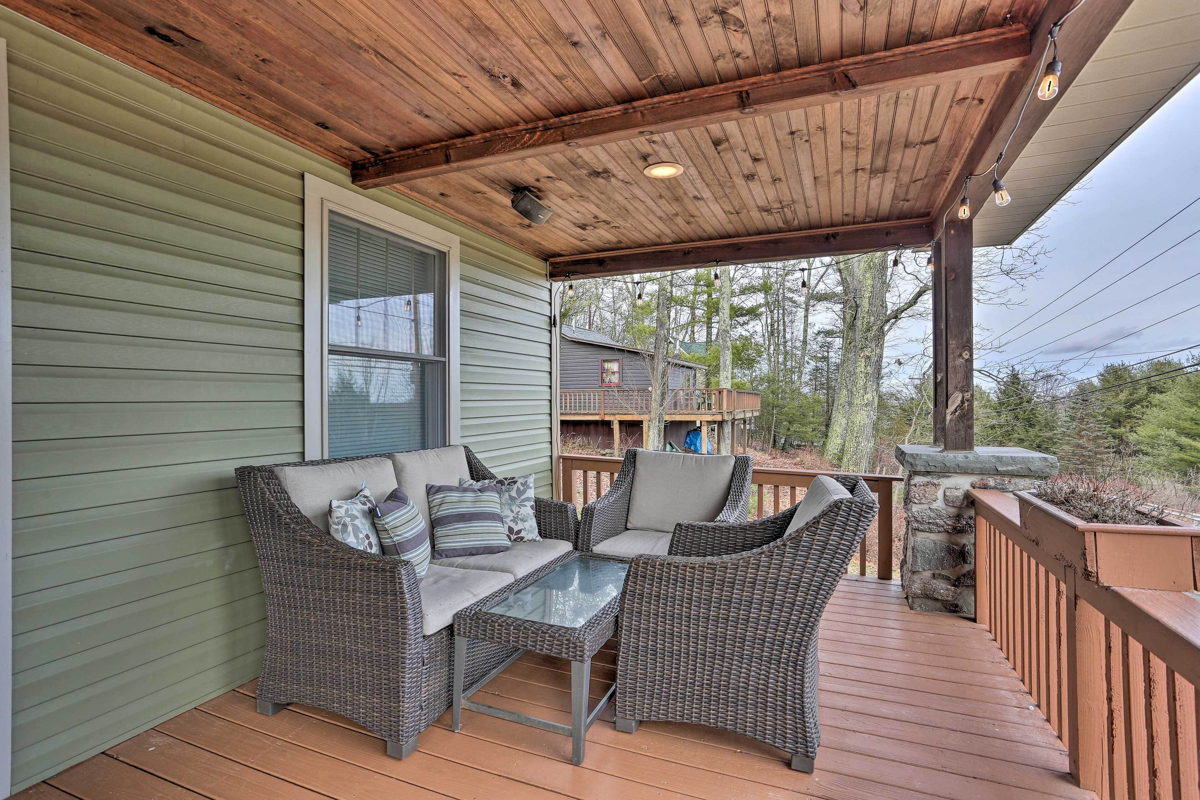 Une véranda aménagée avec des meubles en osier et une table en verre, donnant sur une terrasse en bois et une maison dont la façade est agrémentée de pierres apparentes.