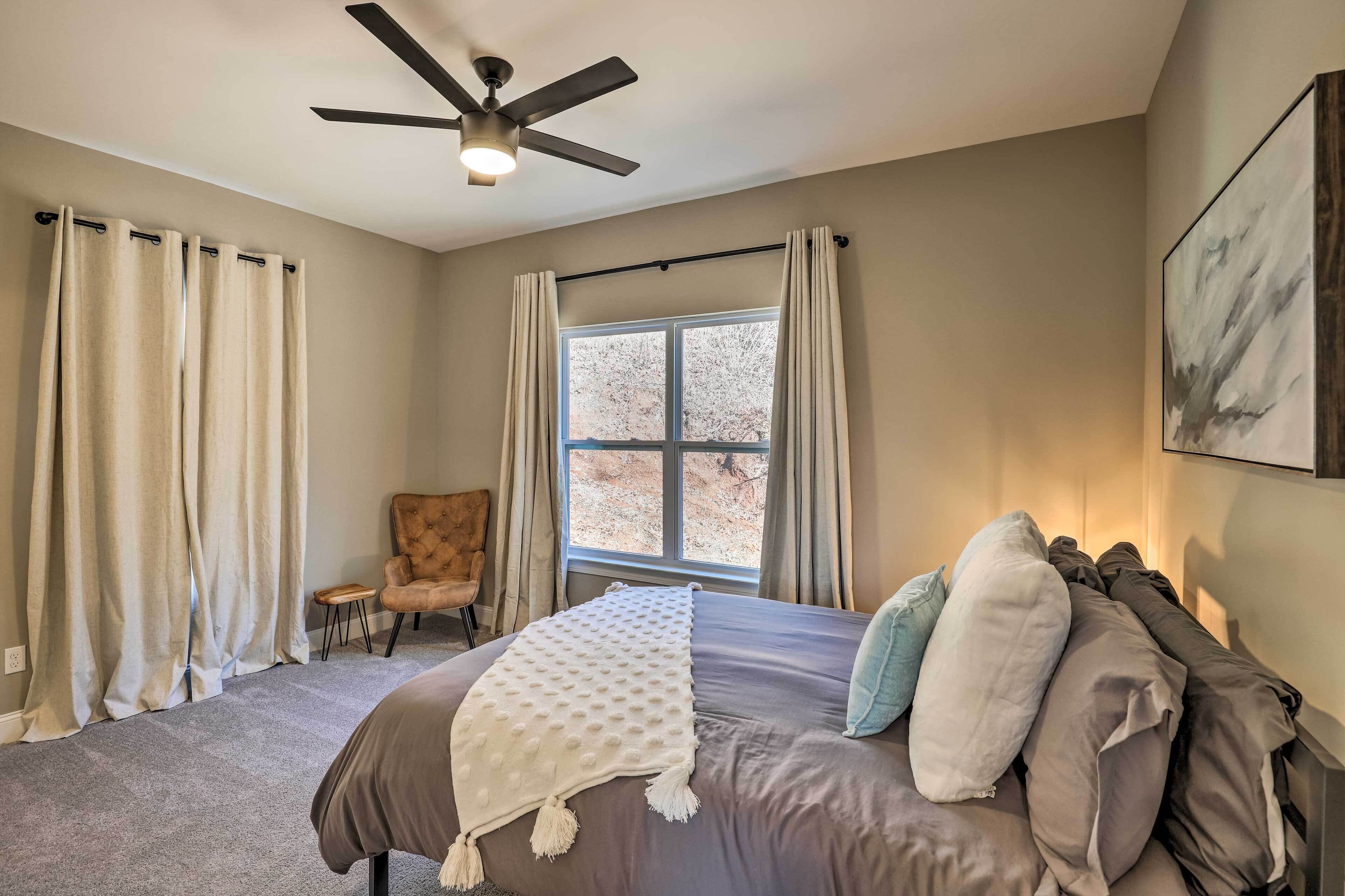 A bedroom with a bed, a chair, a window with curtains, a ceiling fan, and a painting on the wall.