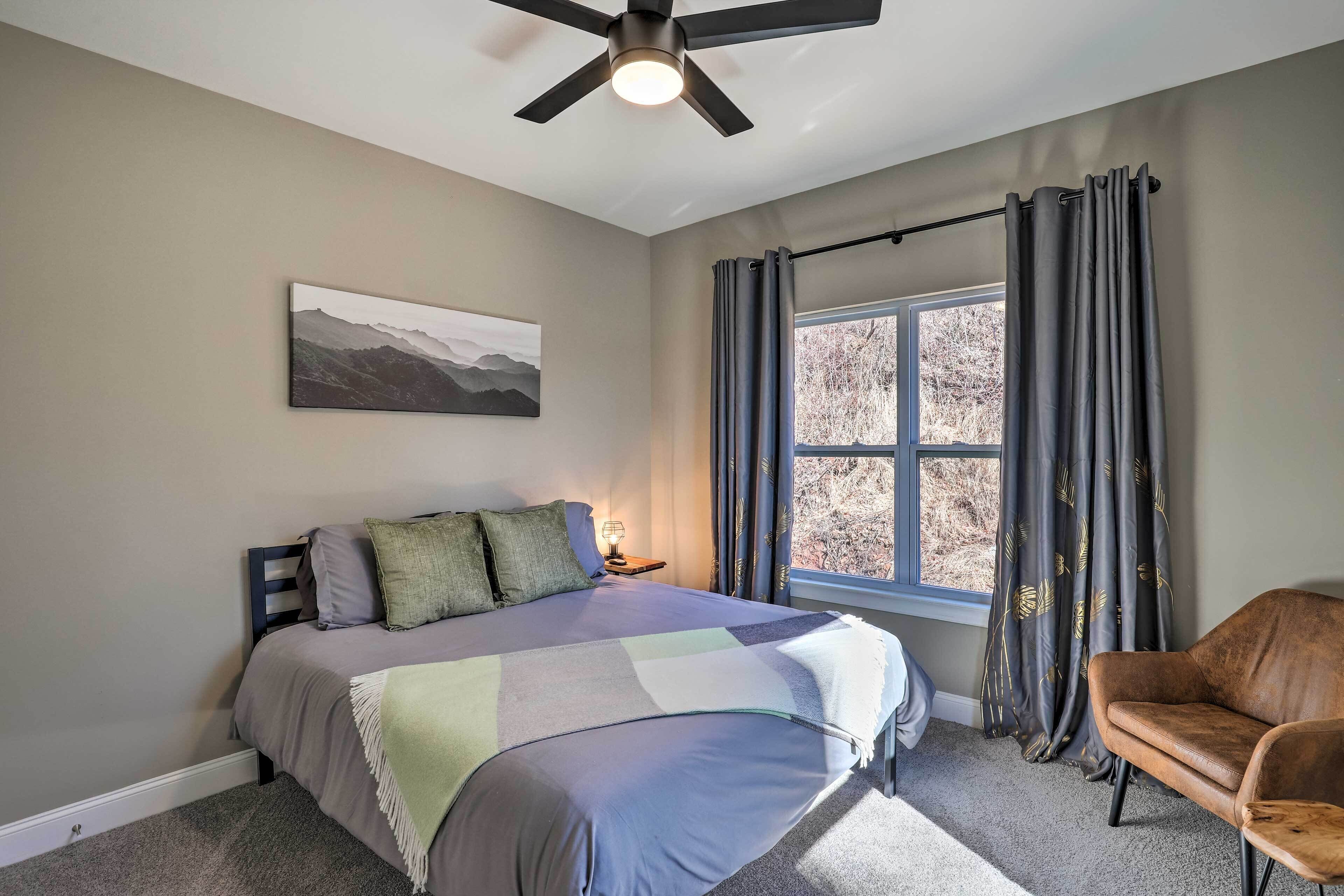 A bedroom with a bed, a chair, a ceiling fan, a window with curtains, and a painting on the wall.