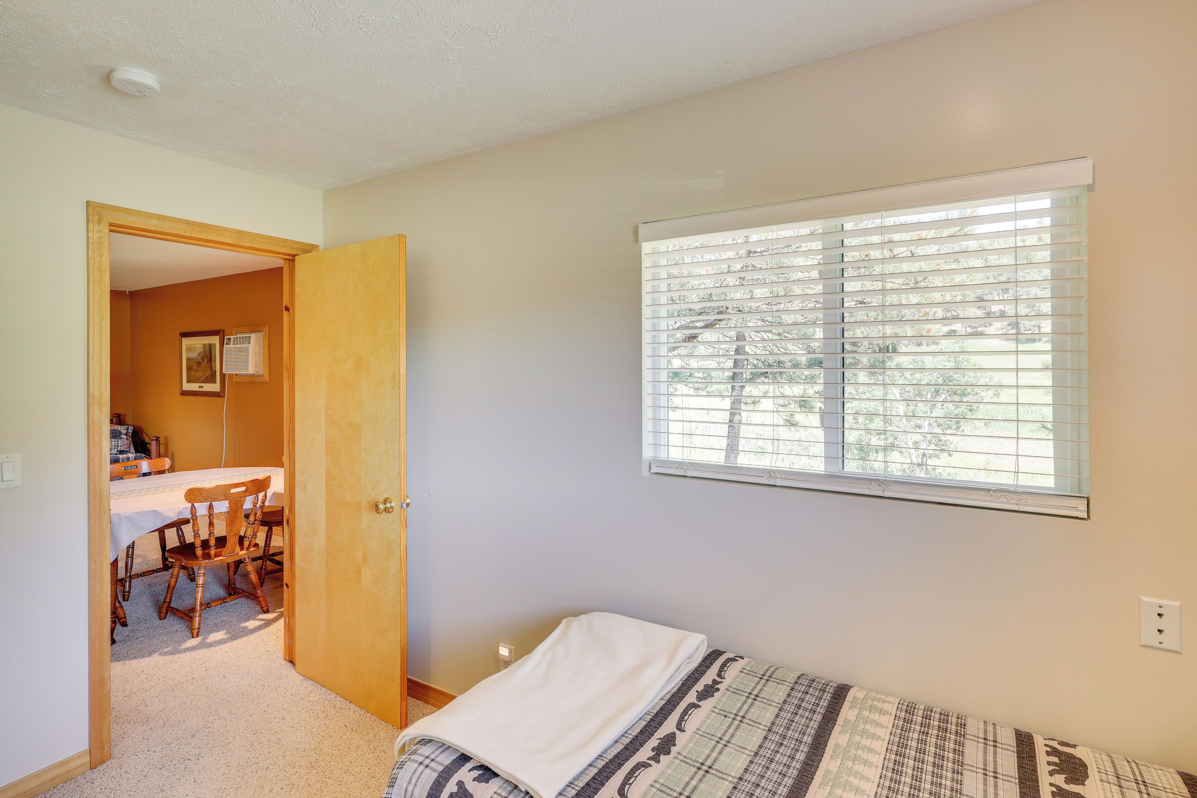 A bedroom with a single bed, a window with blinds, a door leading to another room, and a carpeted floor.