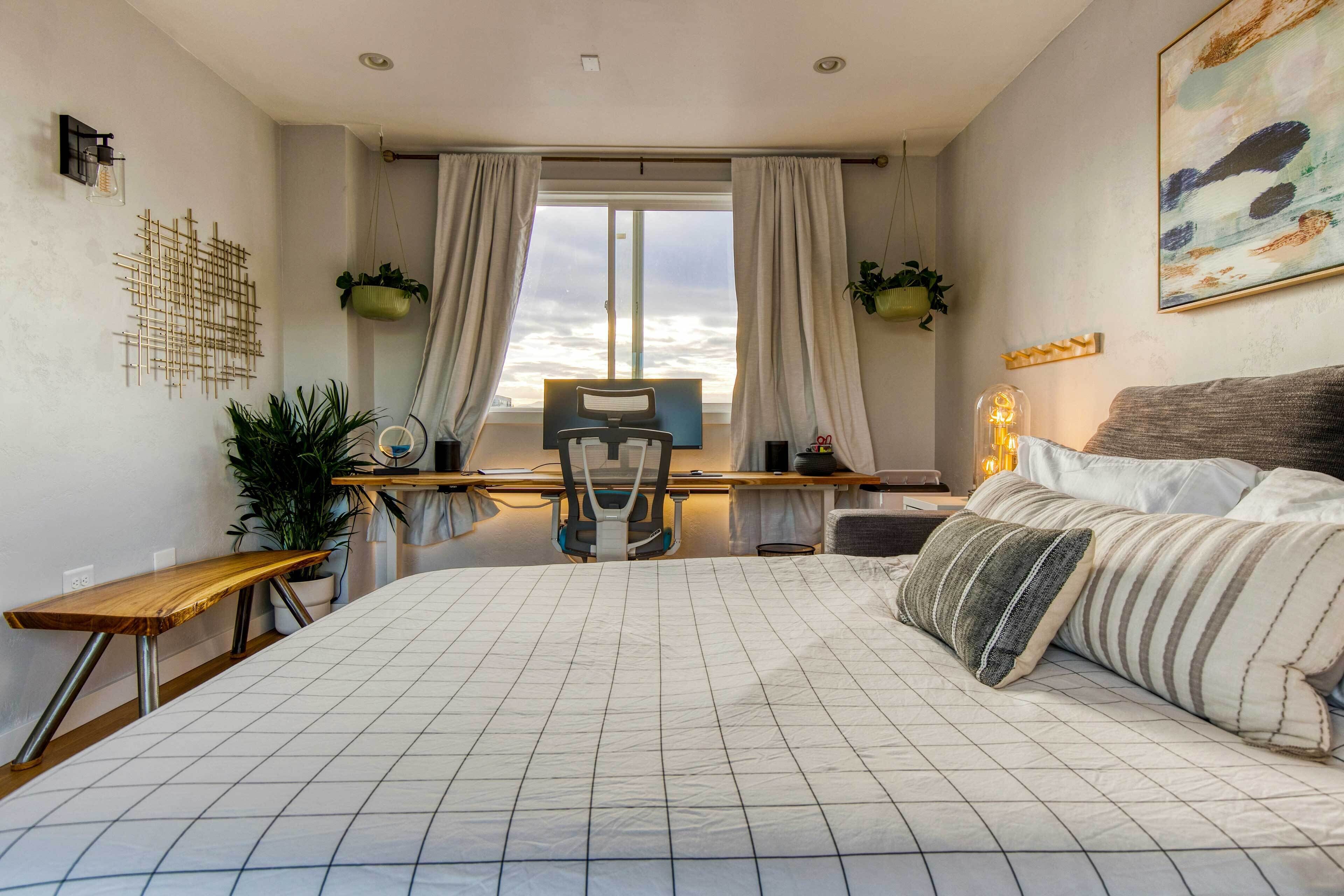 A bedroom with a large bed, a desk, and a chair. There are plants and a painting on the walls.