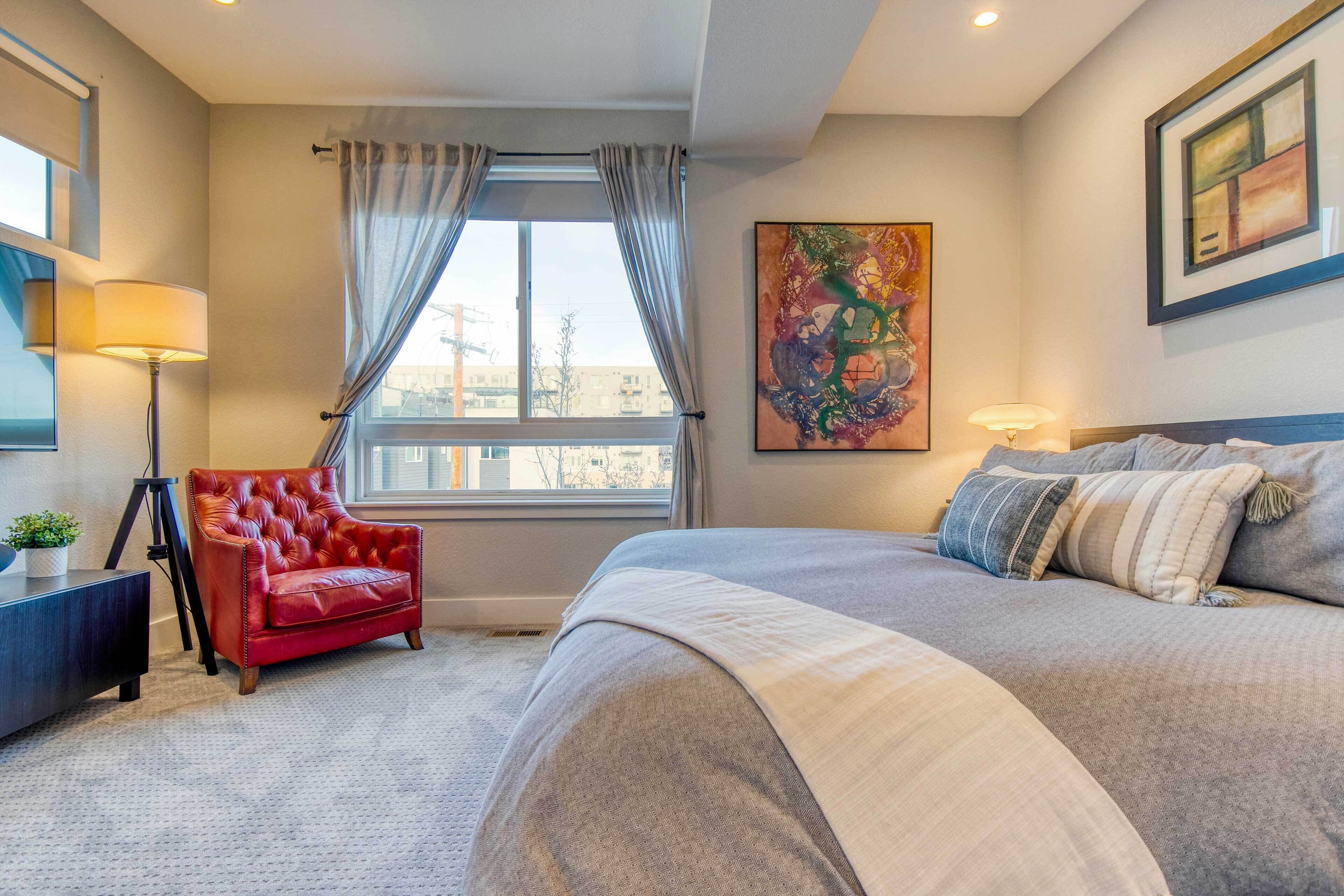 A bedroom with a large bed, a red armchair, a window with curtains, and a painting on the wall.
