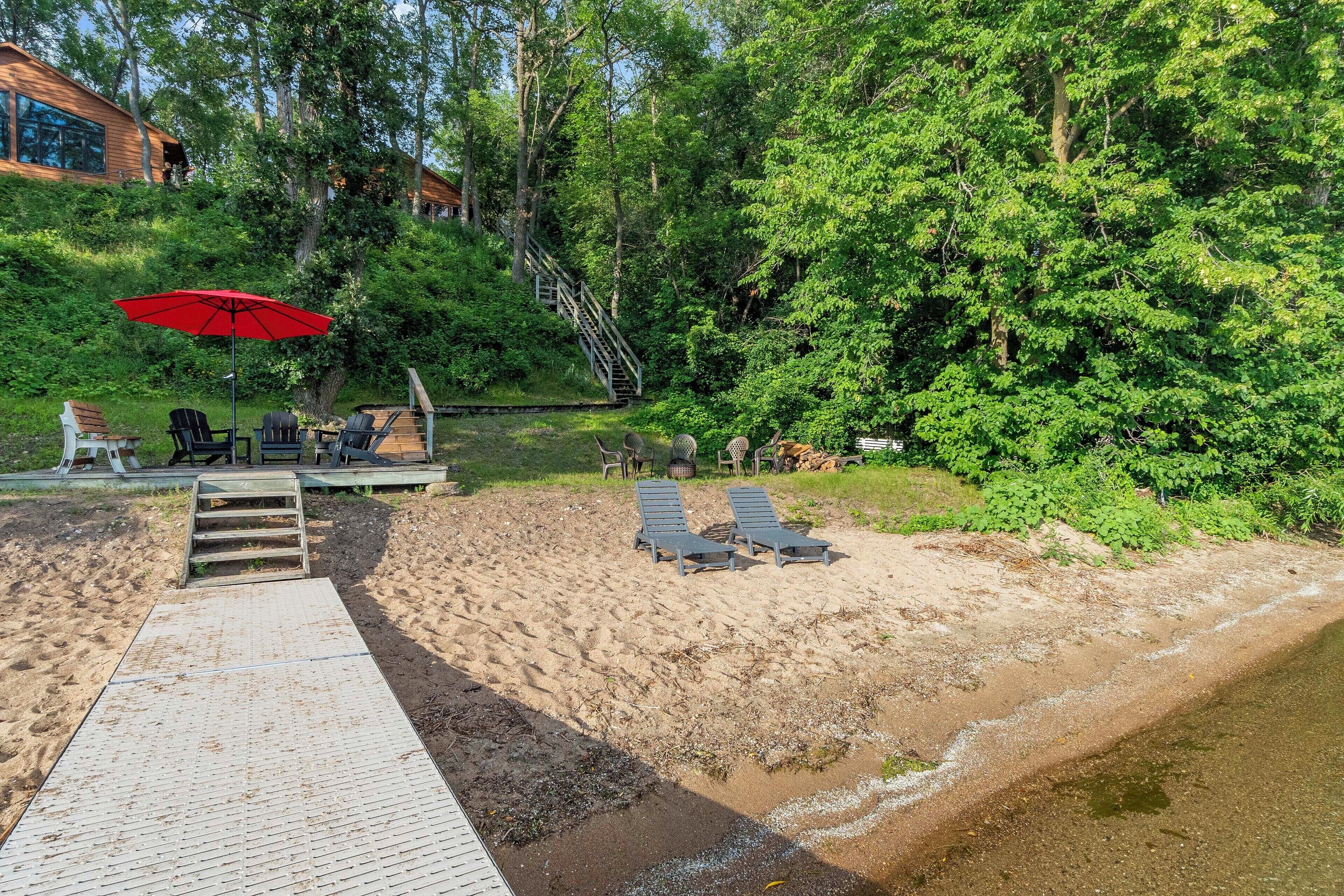 Een terras aan het strand met lounge stoelen en een rode parasol, een houten trap die omhoog leidt, en een zandstrand.