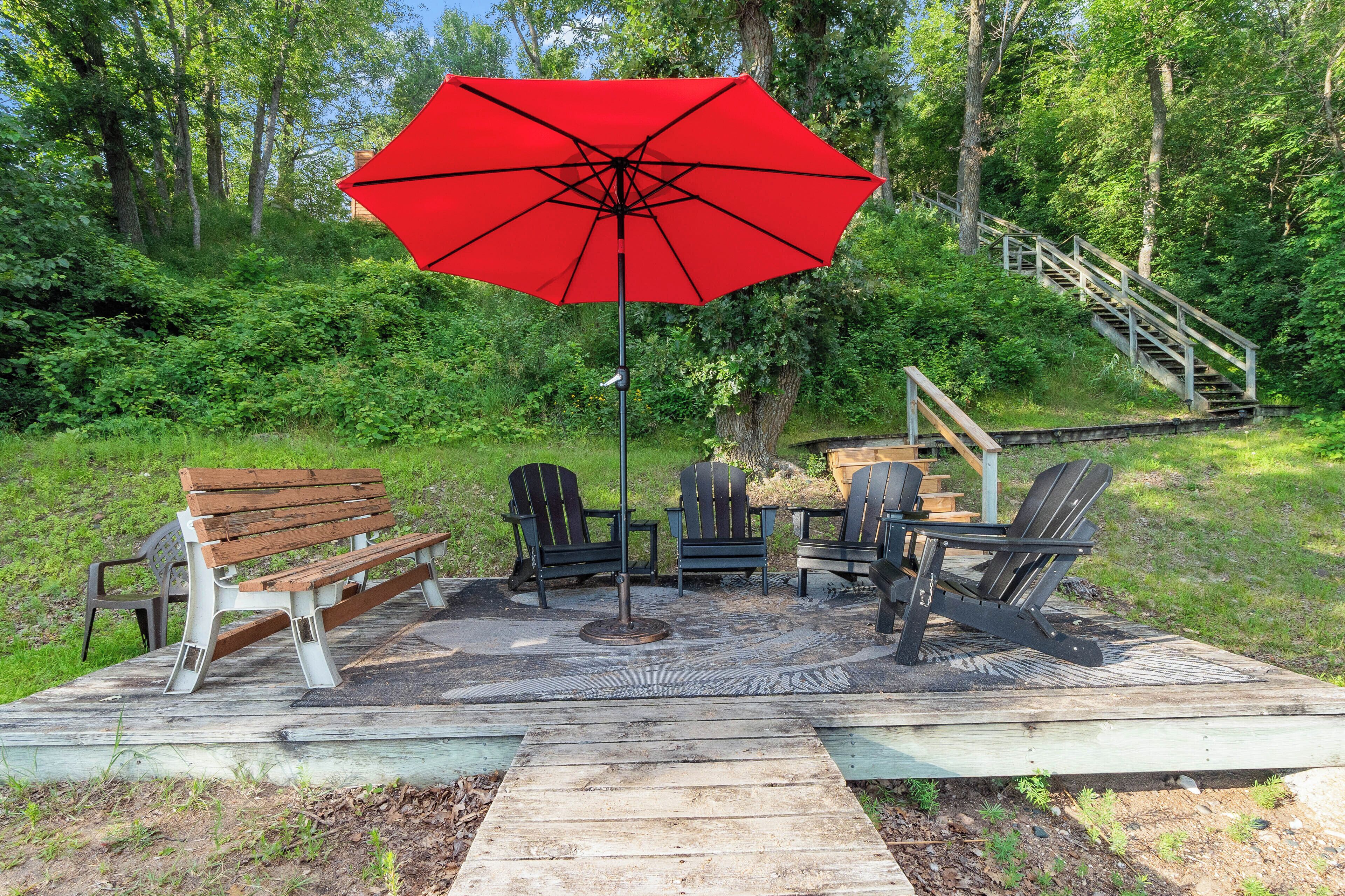 Une terrasse dotée de mobilier d’extérieur, d’un parasol rouge et d’un escalier menant à une zone boisée.