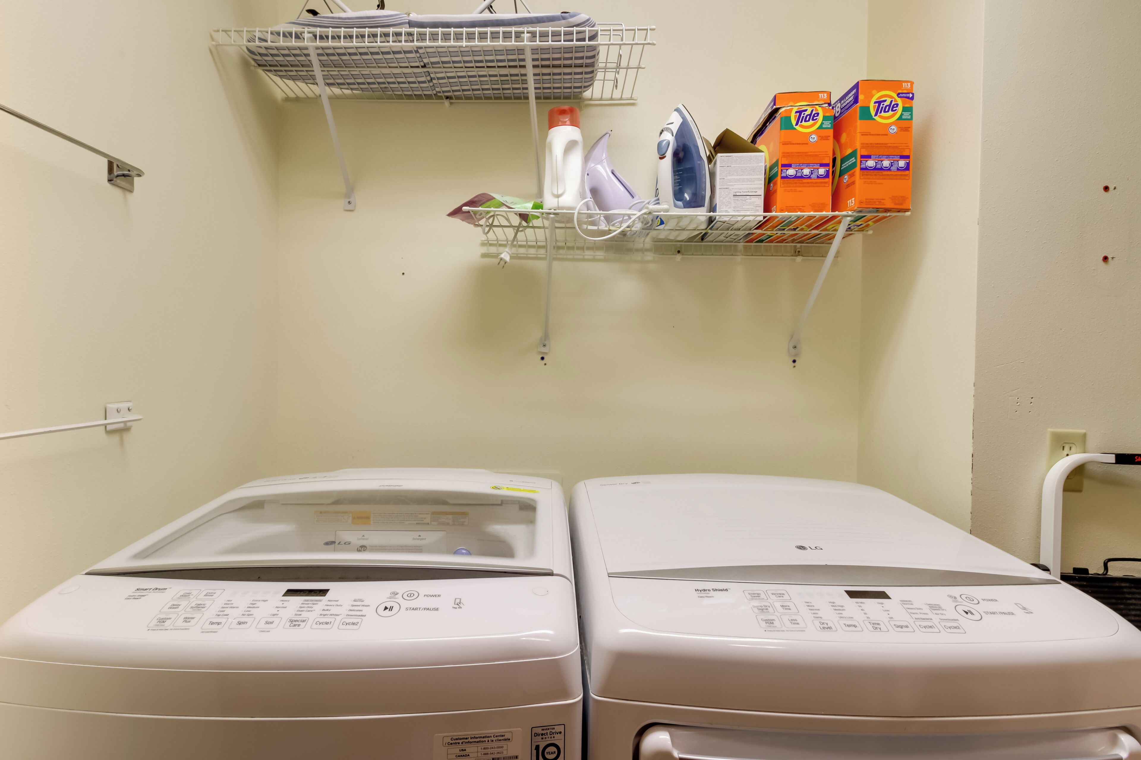 A laundry area with two white LG washing machines, a shelf with laundry supplies, and a hanging rack with folded clothes.