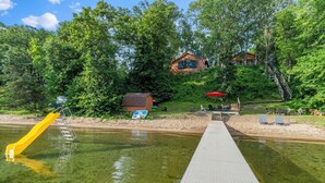 A wooden house surrounded by trees, a dock extending into a clear body of water, and a yellow slide. - Sandy Beach Family Getaway w/ Fire Pit & Deck! (Alexandria)