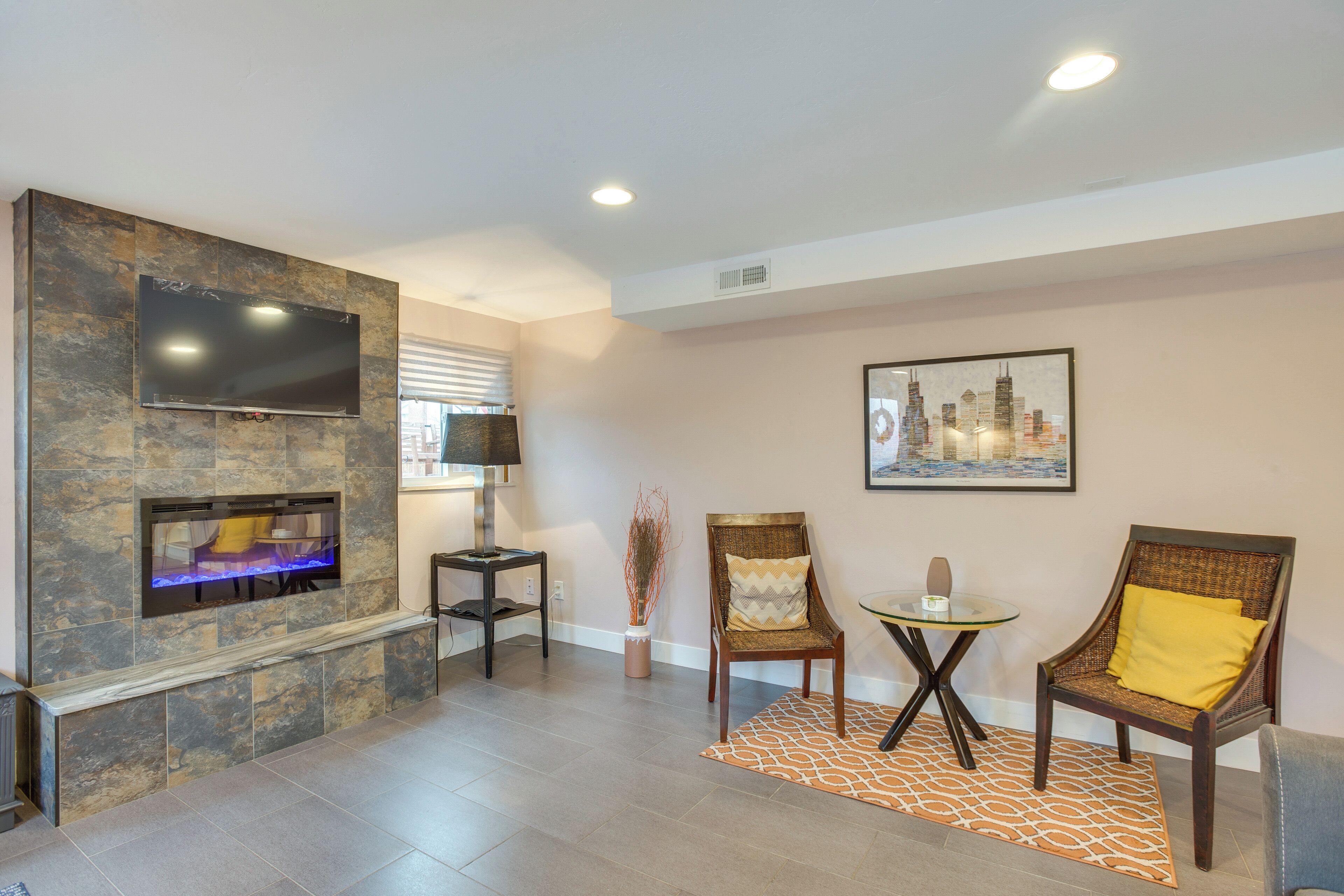 Una sala de estar con chimenea de piedra, un televisor de pantalla plana, dos sillones de mimbre, una mesa de vidrio y un cuadro enmarcado con una vista urbana.