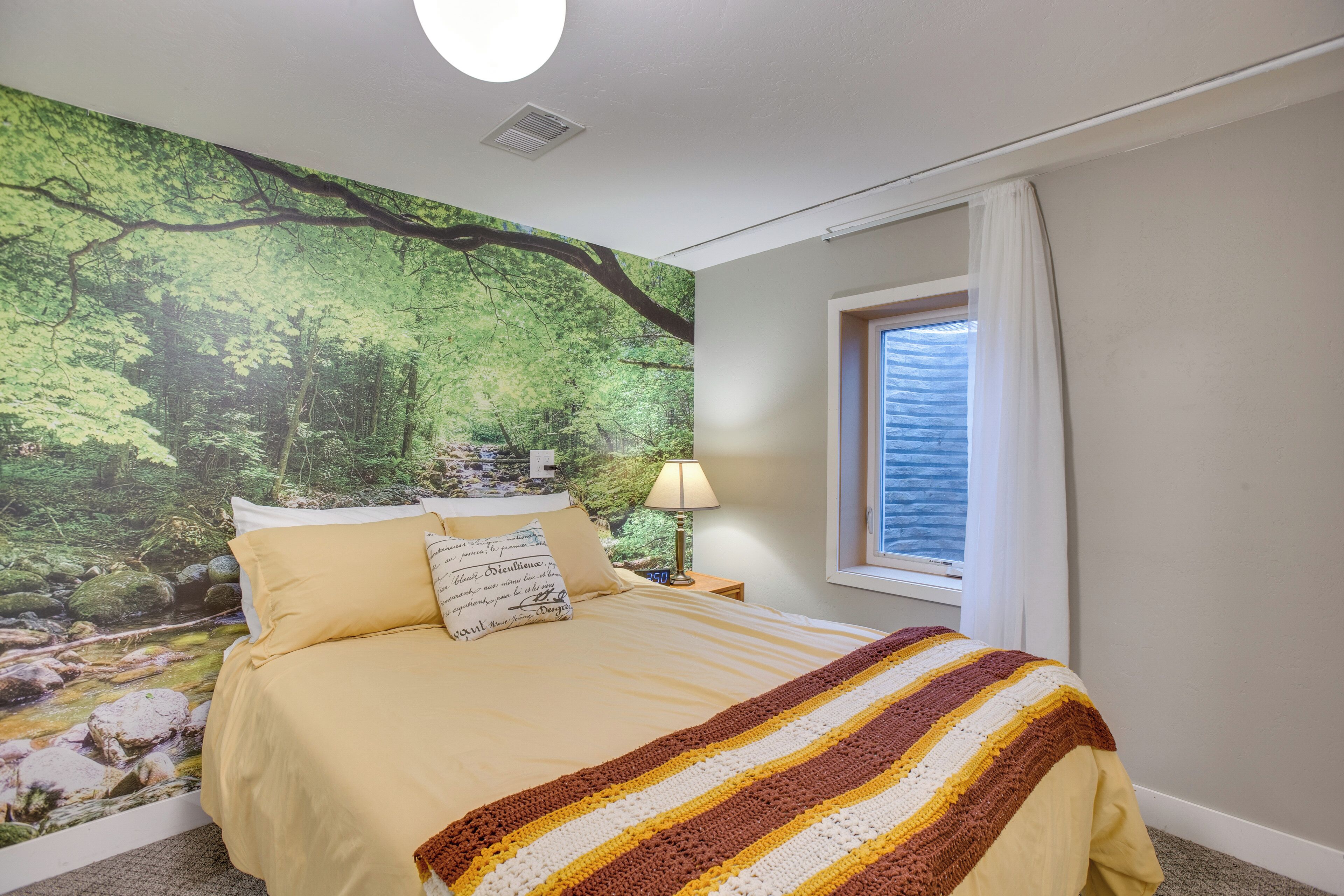 A bedroom with a bed, a nightstand, a lamp, a window with curtains, and a forest mural on the wall.