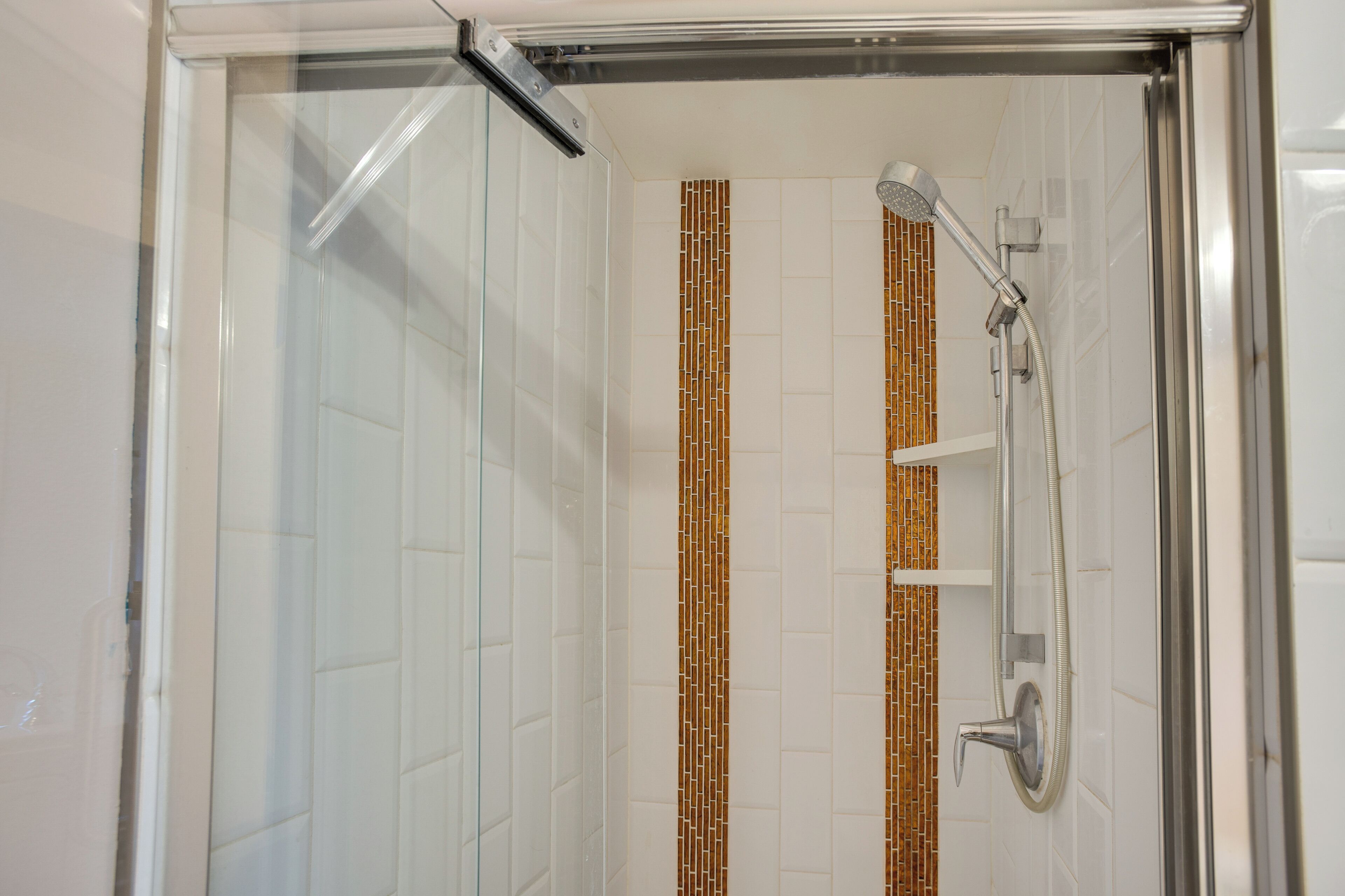 Box de chuveiro com divisória de vidro, chuveiro e mangueira, paredes com azulejos brancos e faixa decorativa com azulejos marrons e brancos.
