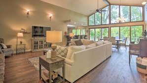 A spacious living room with a large sofa, a television, and a dining area with natural light from large windows. - Lakefront Home w/ Fishing Dock & Fire Table! (Hollister)