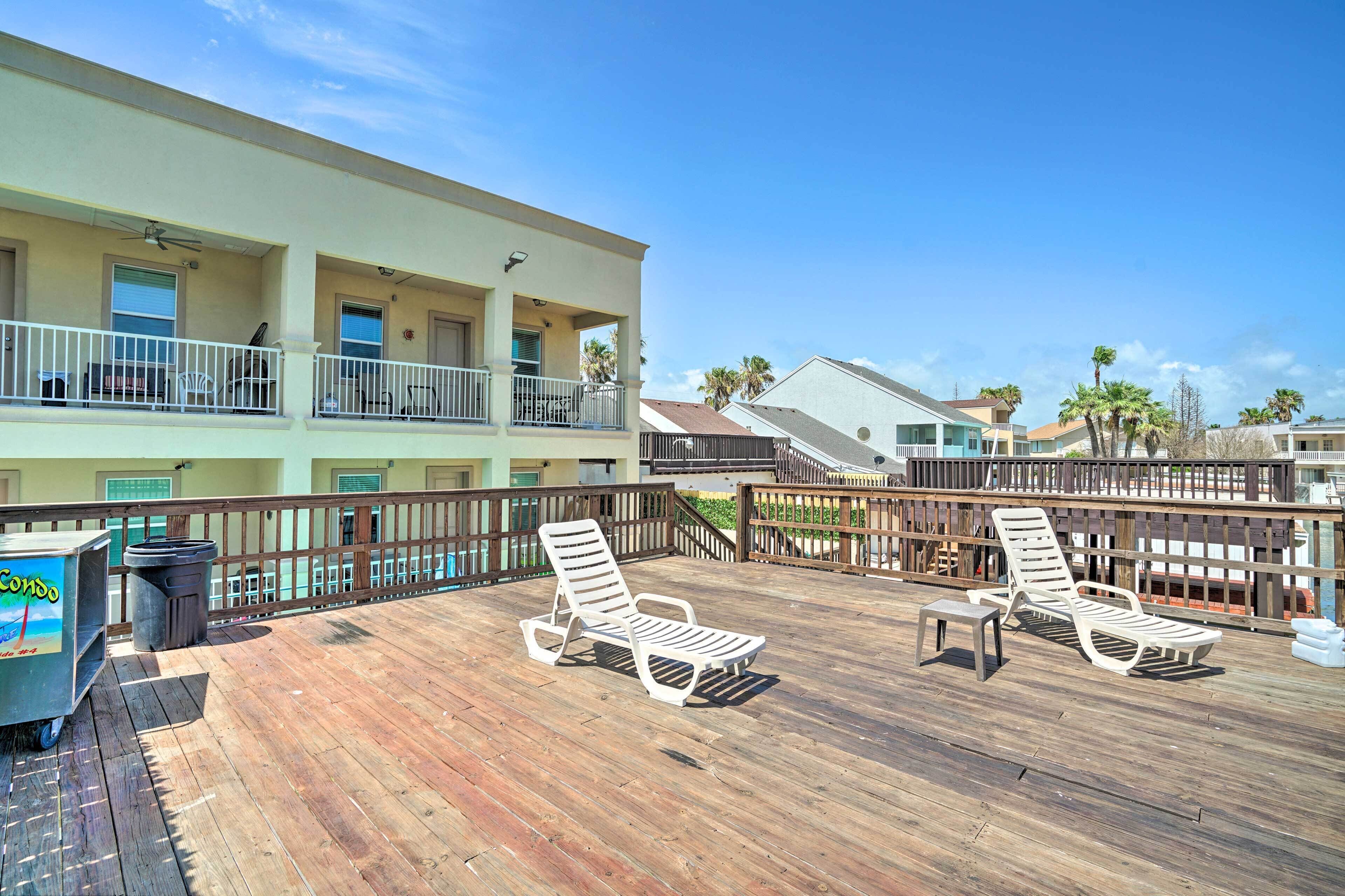 A wooden deck with two sun loungers and a trash bin, overlooking a building with balconies and palm trees.