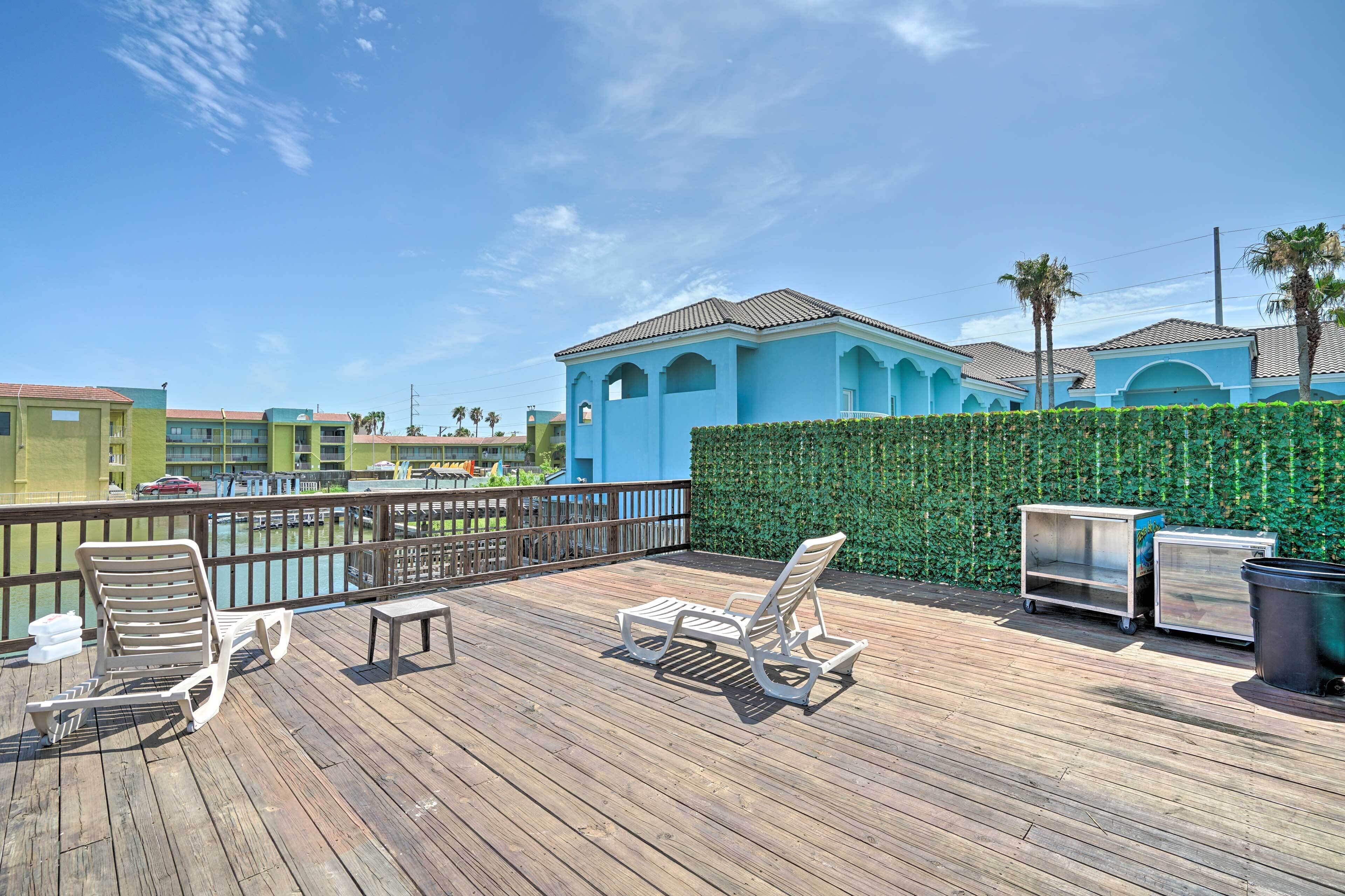 A wooden deck with lounge chairs and a table, a hedge, and a building with a blue facade.