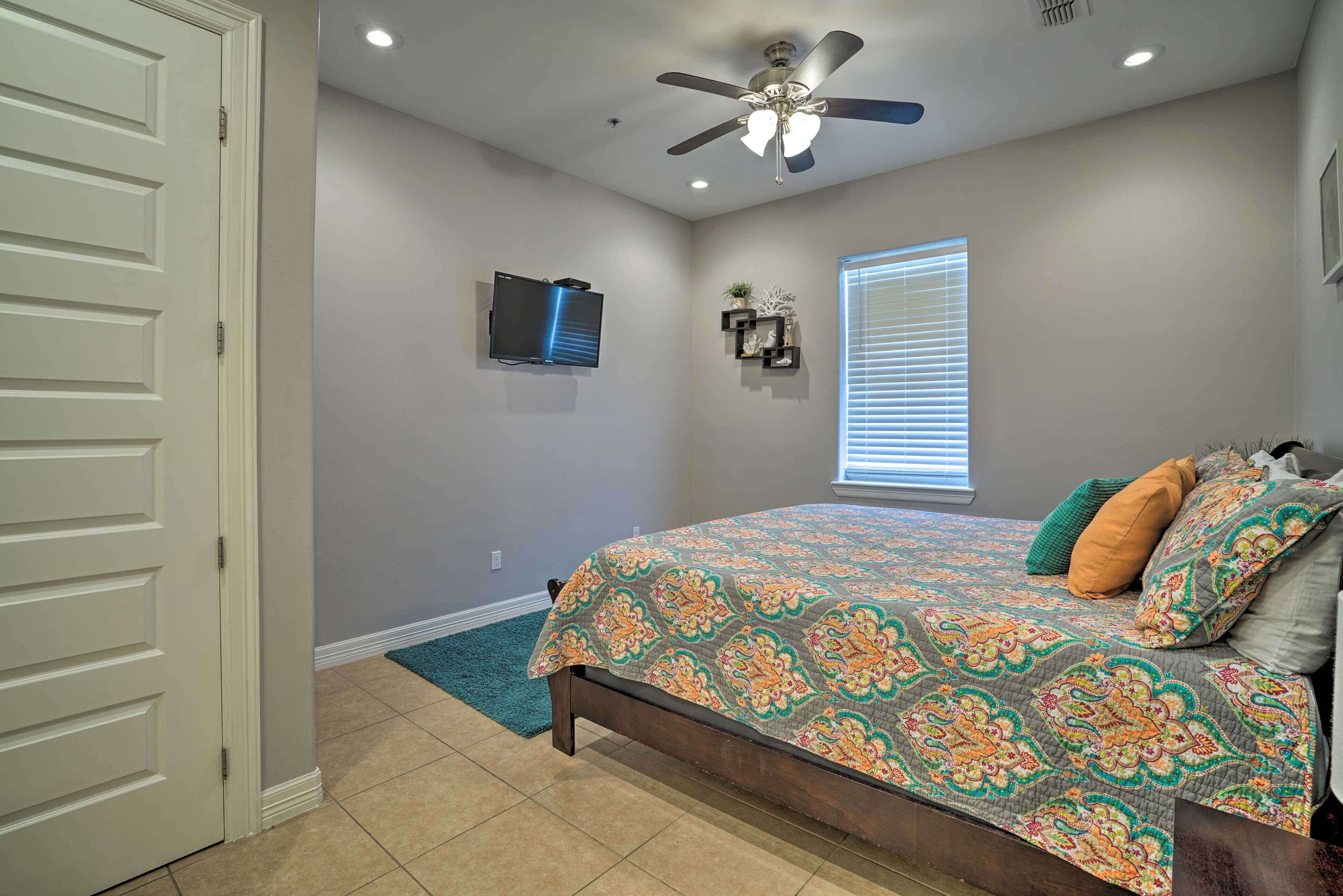 A bedroom with a bed, ceiling fan, TV, window, and a door.