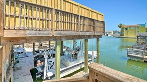A wooden pier with boats docked underneath, a clear sky, and a distant view of a green building. - Updated Fiesta Isles Condo w/ Bay Views & Pool! (South Padre Island)