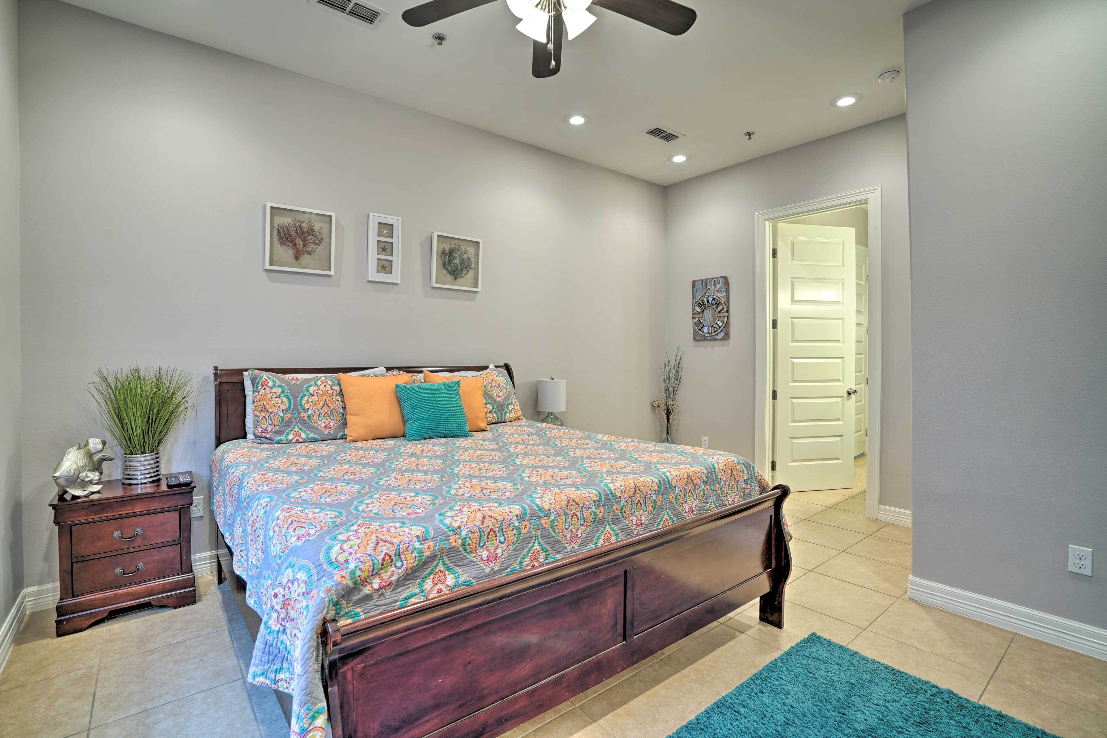 Quarto com cama grande, mesa de cabeceira, ventoinha no teto e porta que dá para outro cômodo.